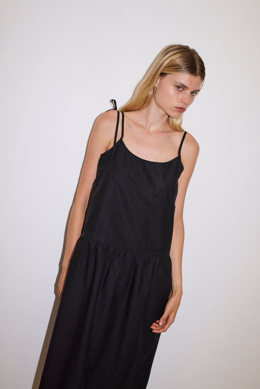 blonde female model wears a black mid-length dress featuring adjustable underlayer straps with sliders for a customised fit and a tied overlayer strap detail at the shoulder