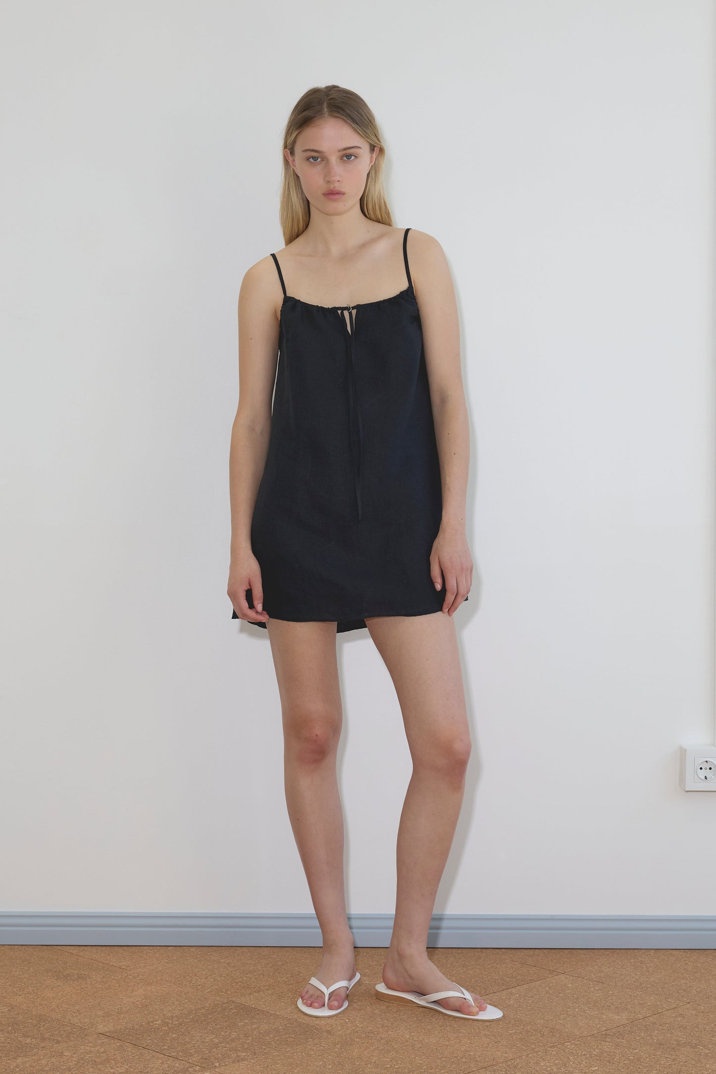A woman with long blonde hair and a neutral expression stands indoors against a white wall, wearing the black Lettuce Mini Dress and white flip-flops.