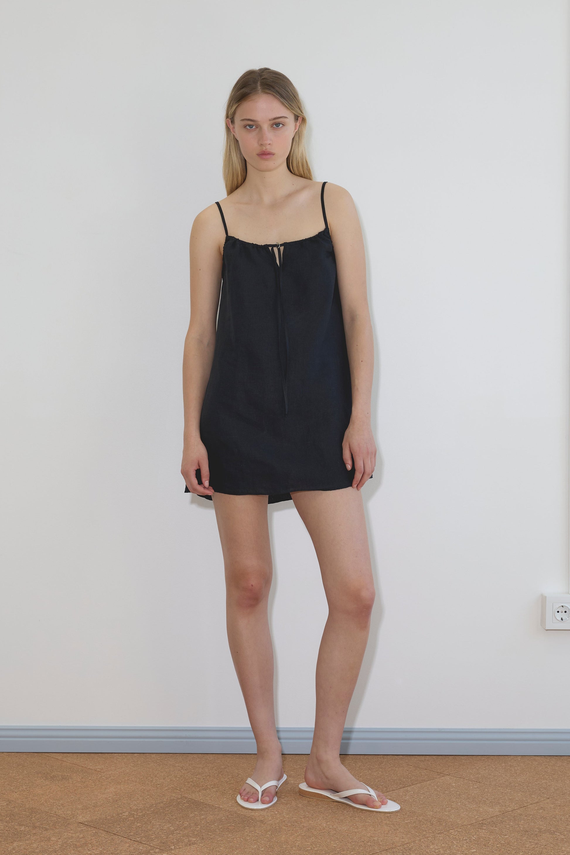 A woman with long blonde hair and a neutral expression stands indoors against a white wall, wearing the black Lettuce Mini Dress and white flip-flops.