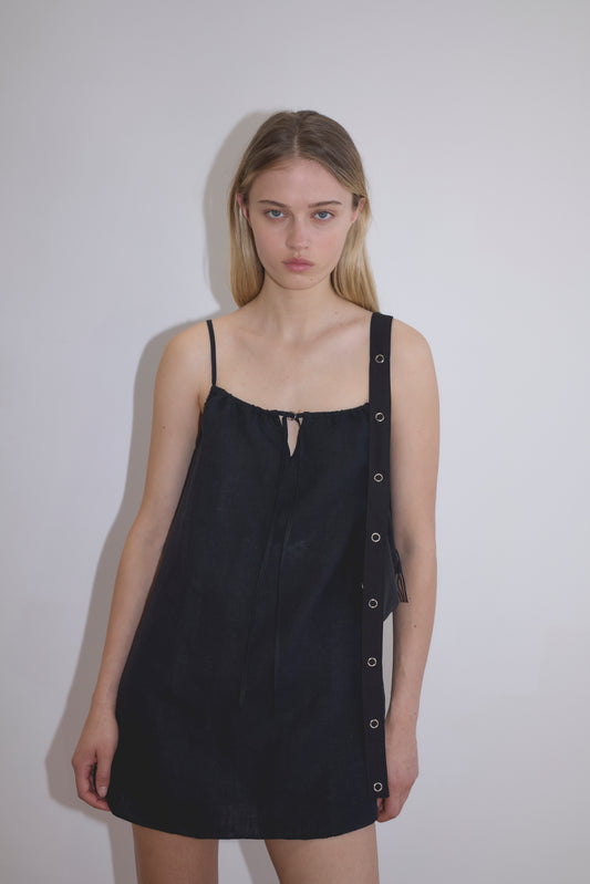 A young woman with straight blonde hair stands against a white wall, wearing the Lettuce Mini Dress - Black, a sleeveless French linen dress with thin straps and grommet details on one side, her expression neutral.
