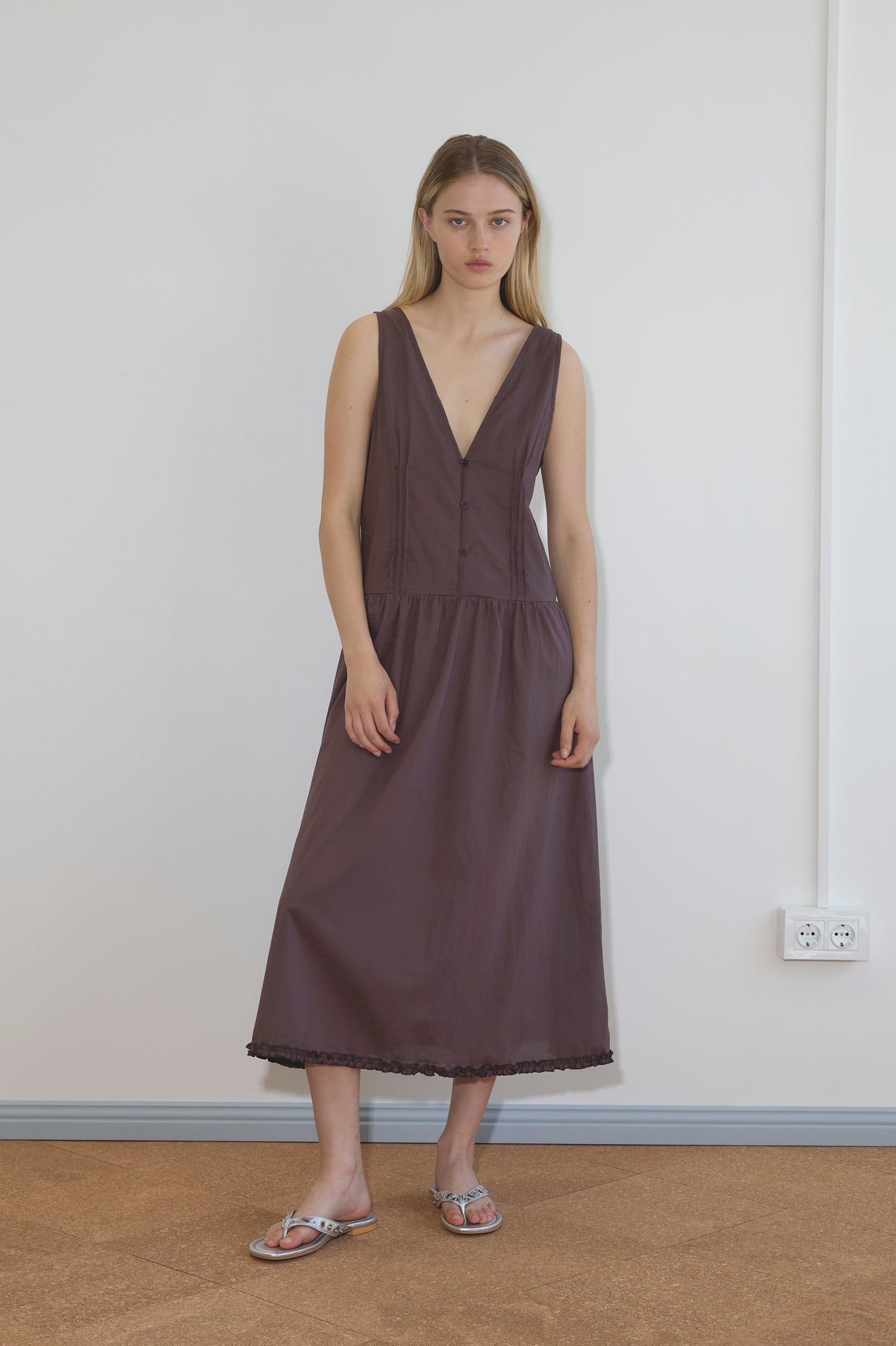 A young woman with long, straight blonde hair wears a sleeveless Deiji Studios Pintuck Dress – Raisin in organic cotton, falling below the knees. She stands indoors on a brown floor against a white wall, looking at the camera.