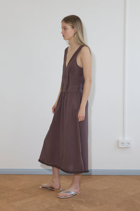 A woman stands indoors against a plain white wall, wearing Deiji Studios' sleeveless Pintuck Dress - Raisin, which has a deep V-neck and ankle length. She faces left with a neutral expression. A colorful pair of flat sandals and an electrical outlet are visible.