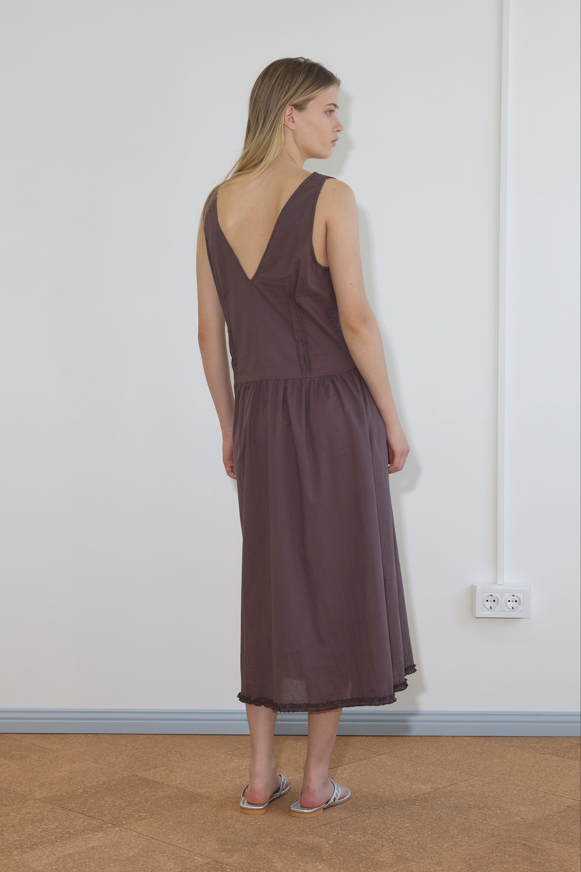 A woman indoors, facing away, wears the Pintuck Dress - Raisin by Deiji Studios—sleeveless, deep V-back, midi-length—and silver flat sandals. She stands near a white wall with a visible power outlet and cord.
