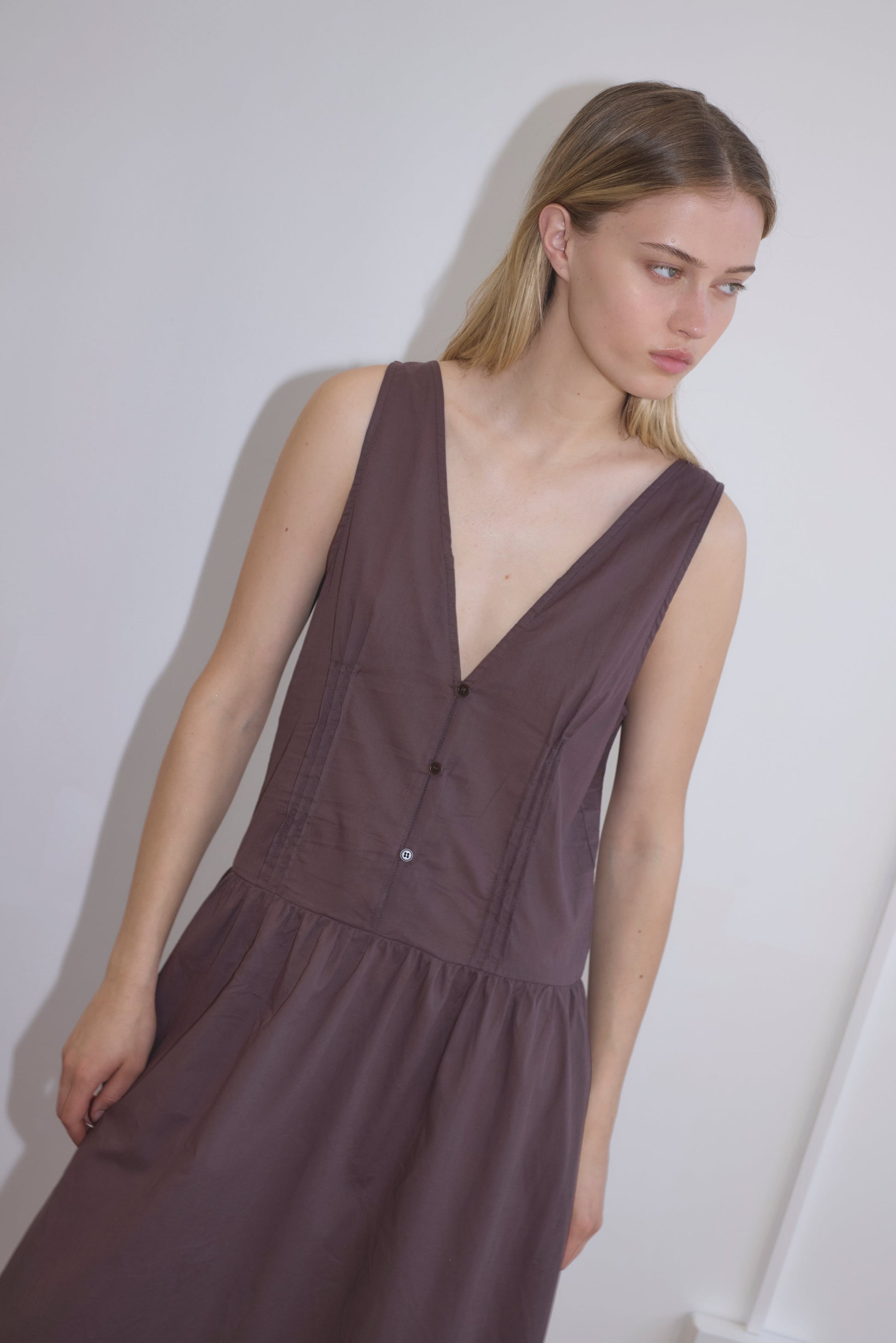 A young woman with straight blonde hair wears the Pintuck Dress - Raisin by Deiji Studios, featuring a sleeveless deep V-neck design, as she stands against a plain white wall and gazes to the side with a neutral expression.