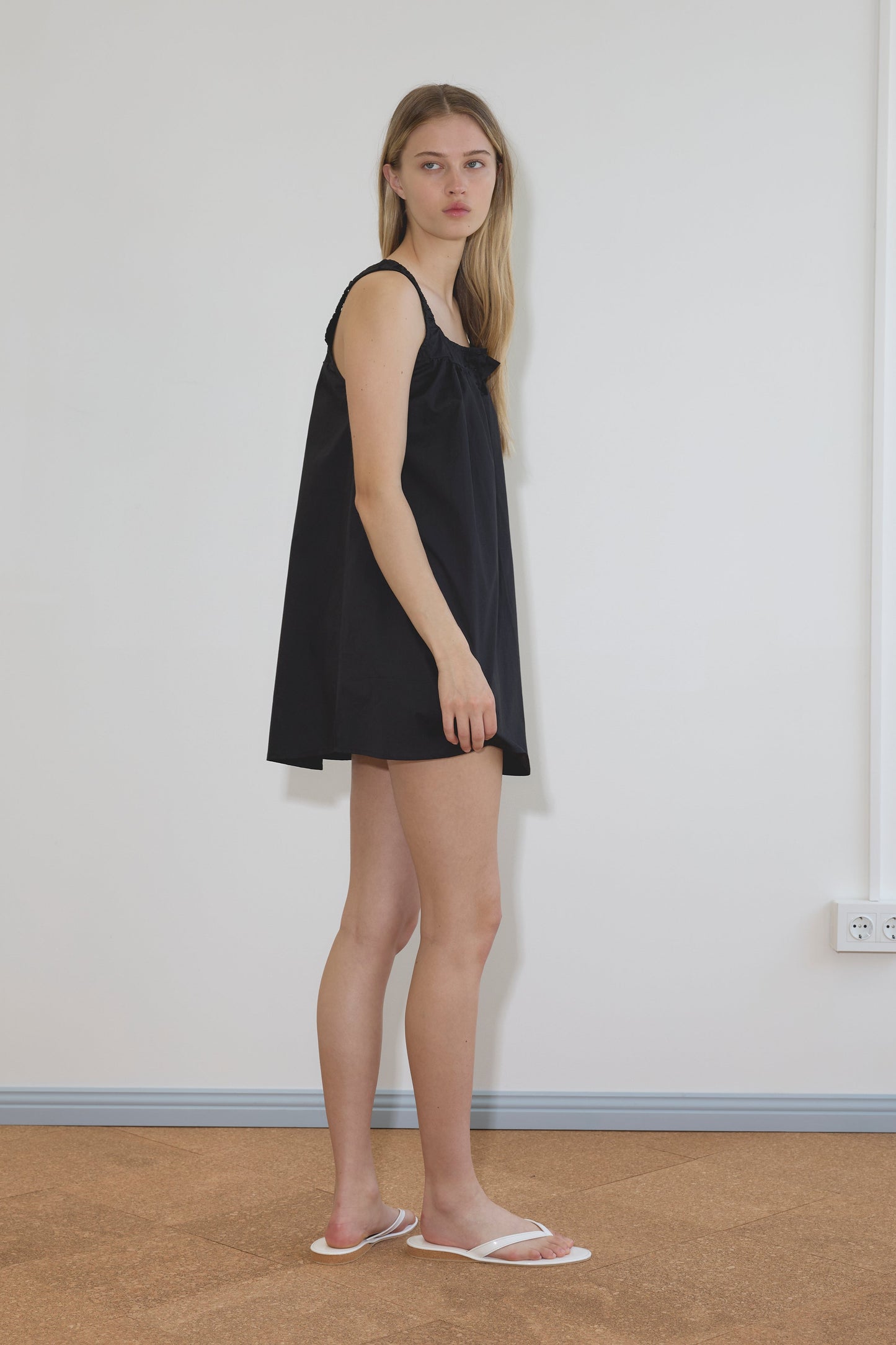 A person with long blonde hair stands sideways against a plain white wall, wearing the Yoke Mini Dress - Black by Deiji Studios and white sandals on a brown floor.