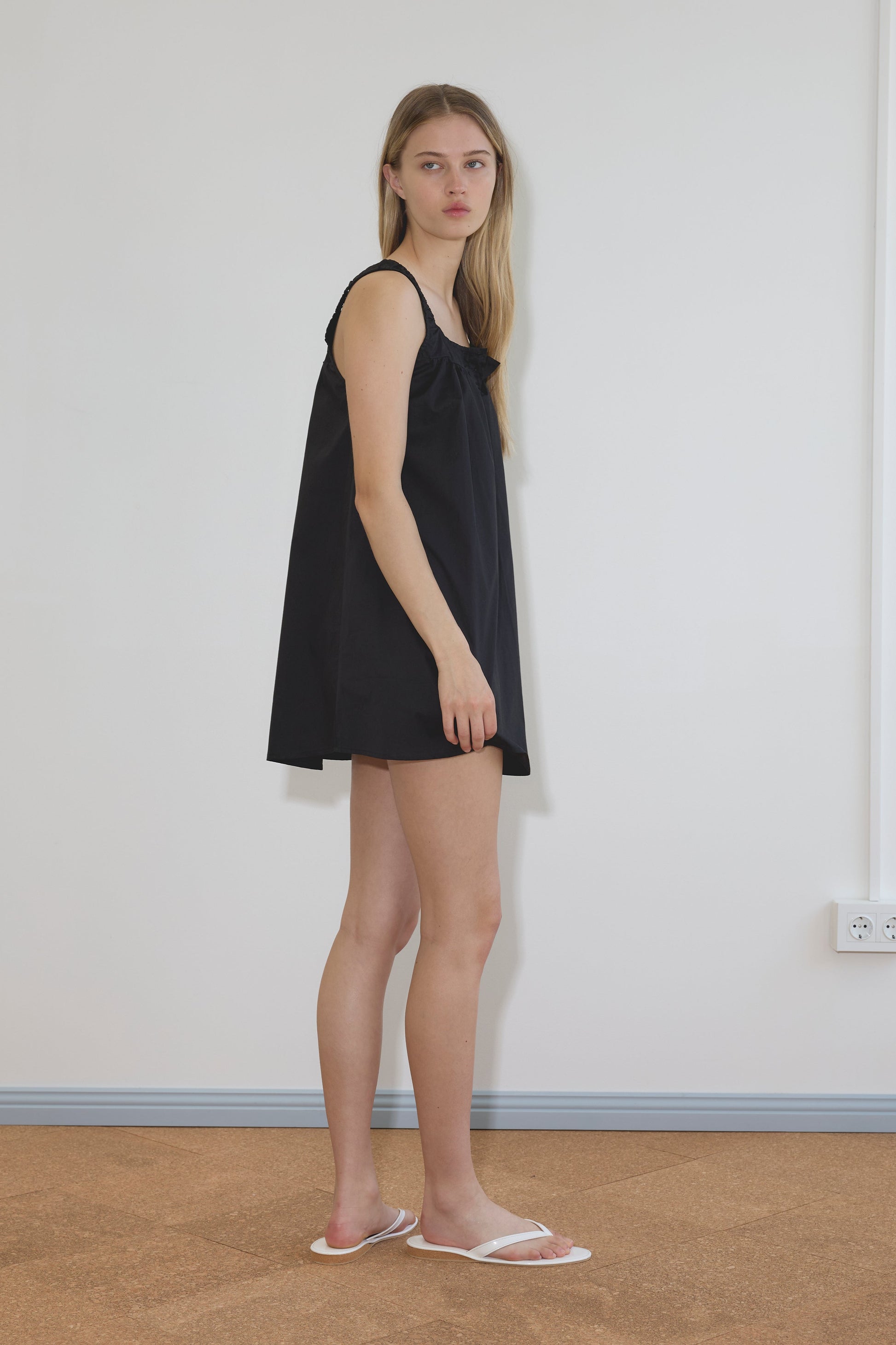 A person with long blonde hair stands sideways against a plain white wall, wearing the Yoke Mini Dress - Black by Deiji Studios and white sandals on a brown floor.