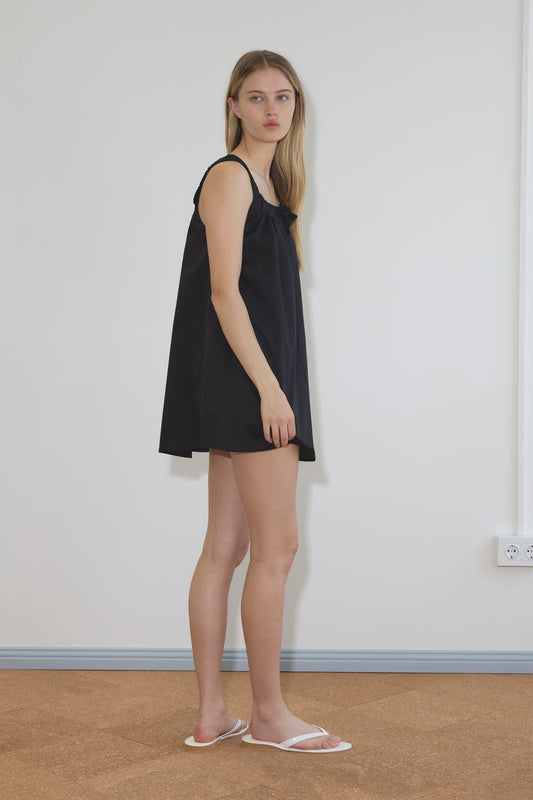 A person with long blonde hair stands sideways against a plain white wall, wearing the Yoke Mini Dress - Black by Deiji Studios and white sandals on a brown floor.