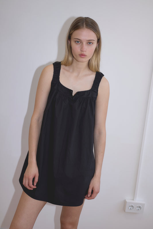 A young woman with straight blonde hair stands against a white wall, wearing the Yoke Mini Dress - Black. She has a neutral expression, her arms relaxed at her sides, and an electrical outlet is visible on the wall.