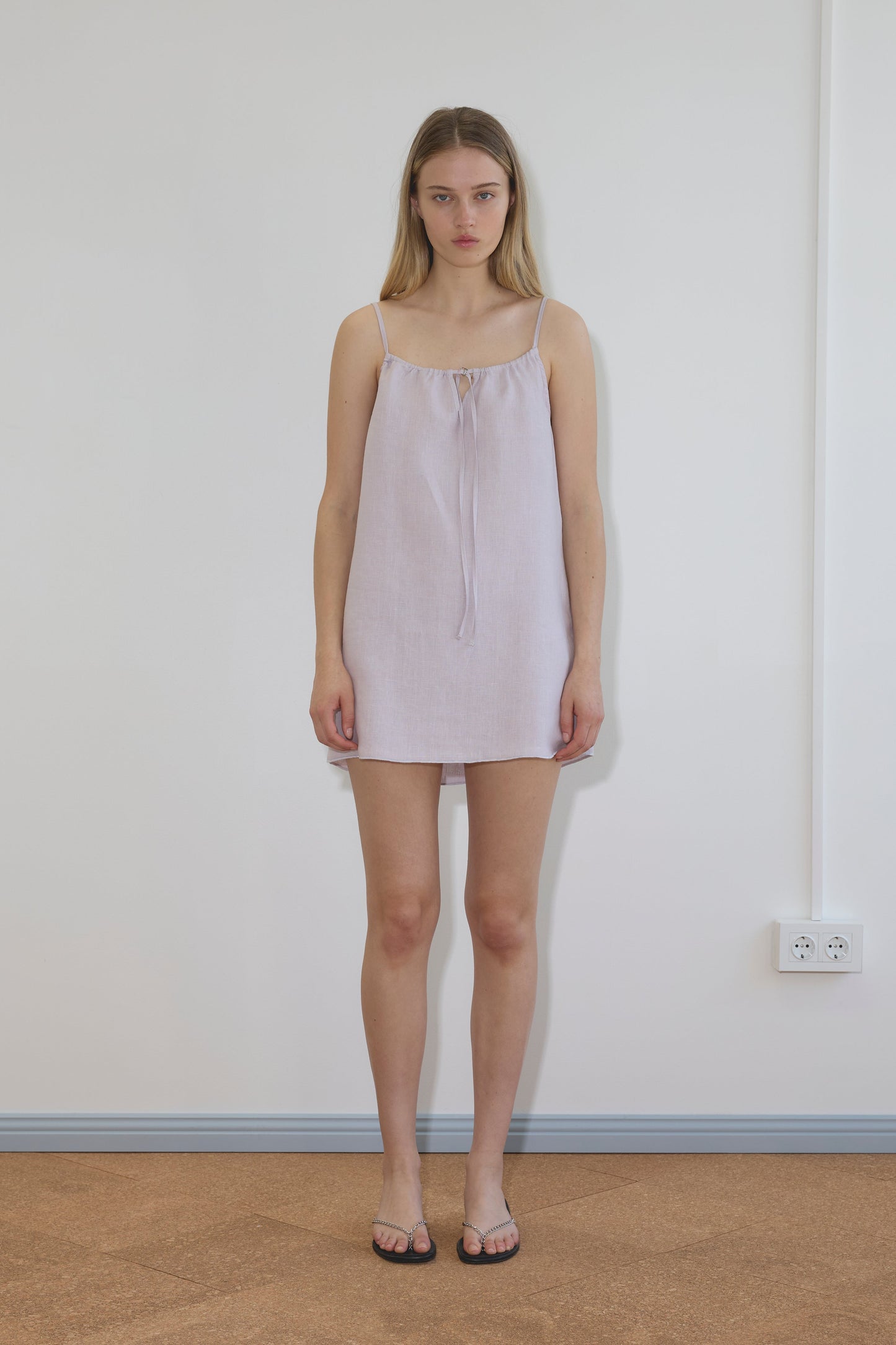 A woman with straight blonde hair stands against a white wall, wearing the Lettuce Mini Dress in Lilac by Deiji Studios—an organic linen dress with thin straps—paired with black sandals, perfectly reflecting the brand's effortless style.