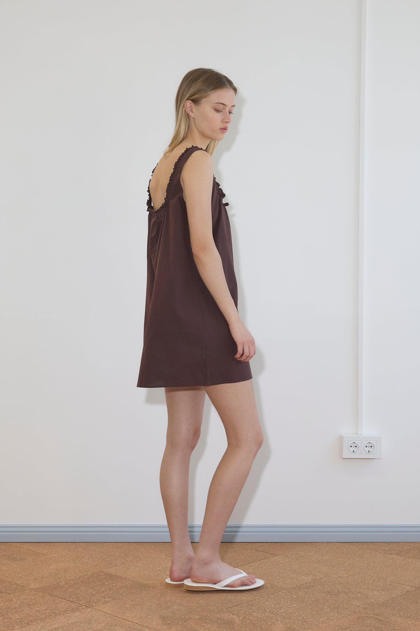 A woman in profile stands against a white wall, wearing the Button Frill Mini Dress - Umber from Deiji Studios—a short, sleeveless brown dress with ruffled straps—and white slide sandals. The tan floor and power outlet emphasize the minimalist scene.