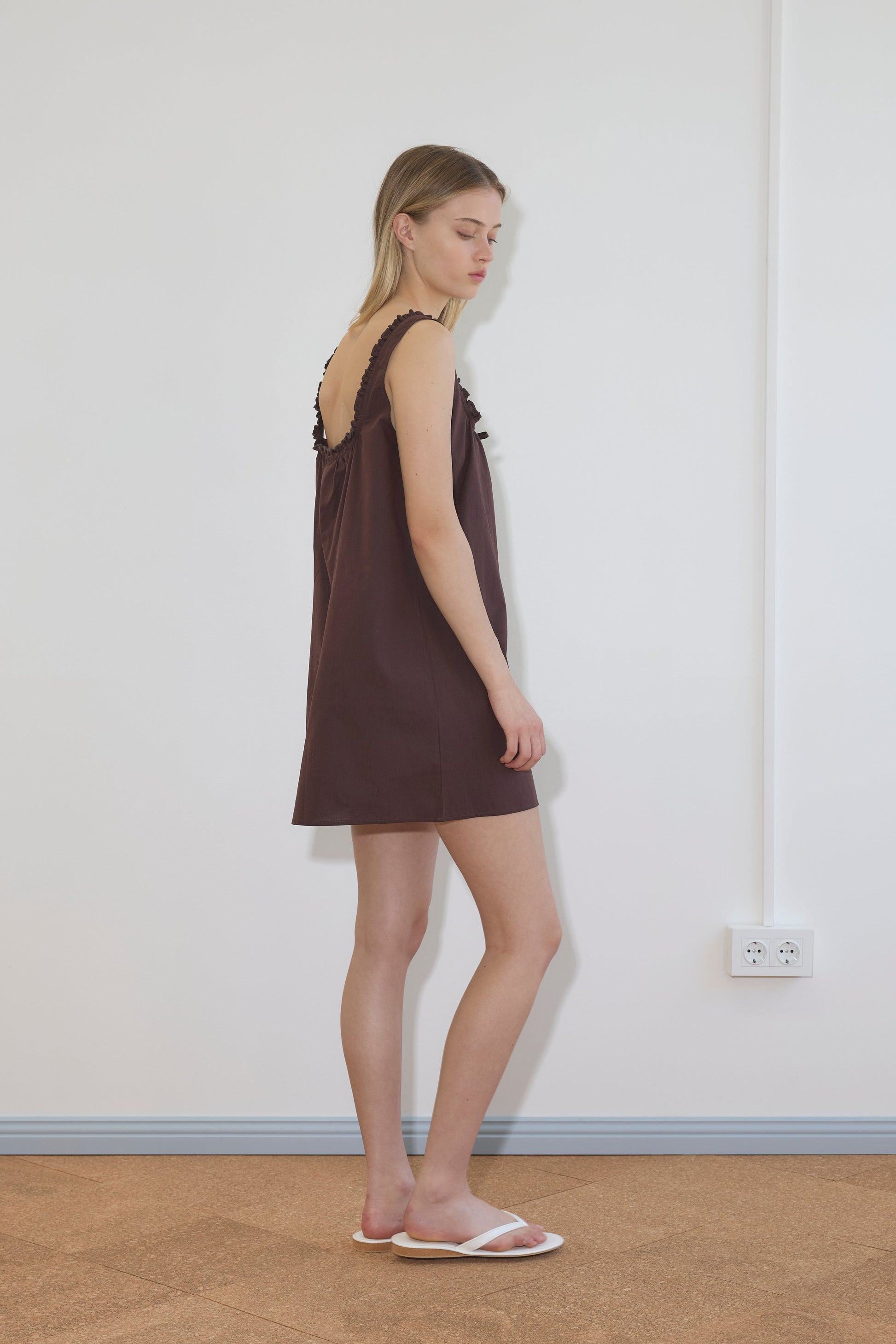 A woman in profile stands against a white wall, wearing the Button Frill Mini Dress - Umber from Deiji Studios—a short, sleeveless brown dress with ruffled straps—and white slide sandals. The tan floor and power outlet emphasize the minimalist scene.