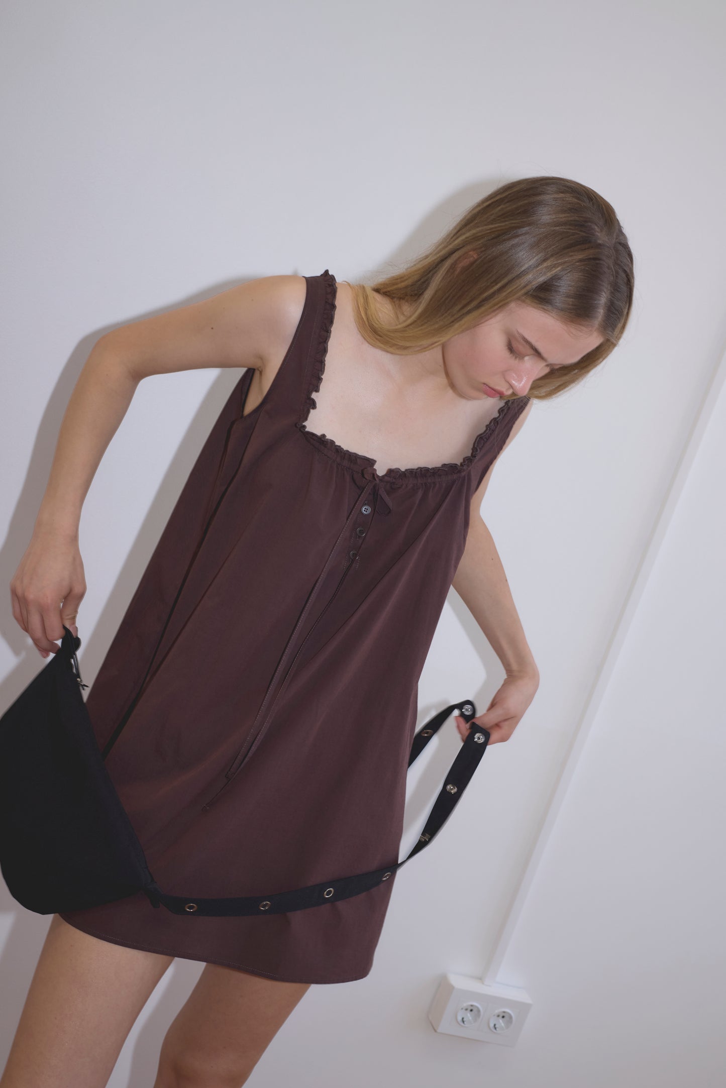 A young woman with straight light brown hair adjusts a black bag strap while wearing the Button Frill Mini Dress - Umber. She stands indoors against a plain white wall near a white power outlet.