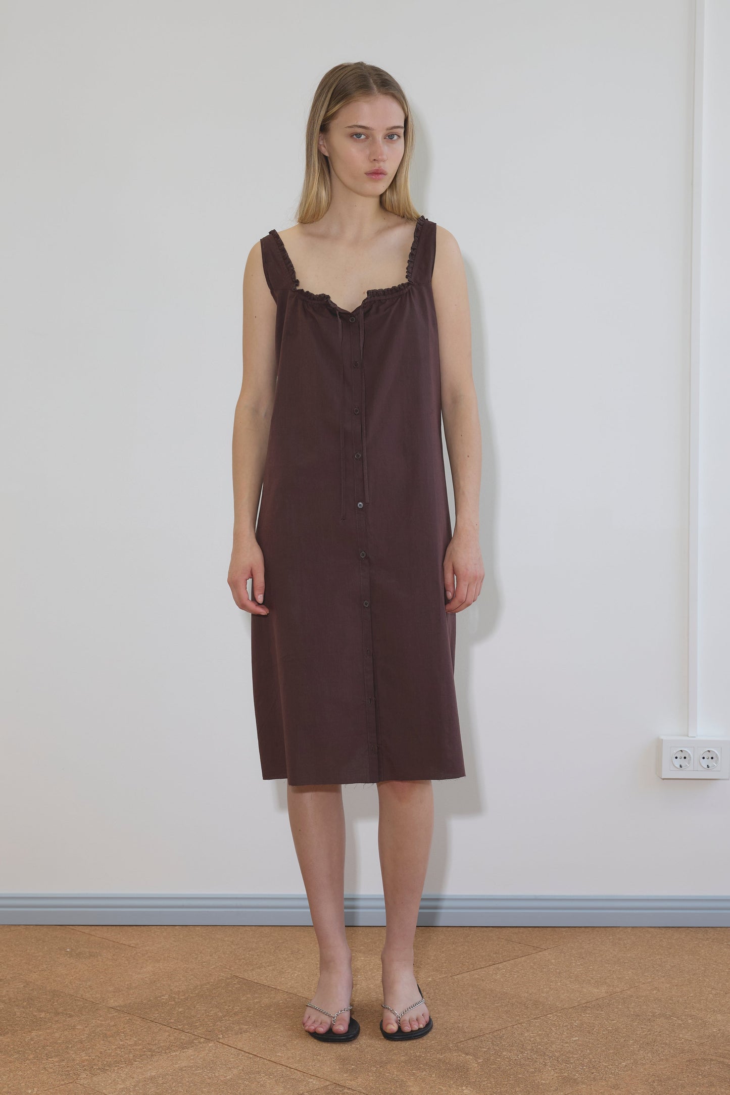 A woman with straight blonde hair stands indoors against a white wall, wearing the Button Sweep Dress - Umber and black sandals. She has a neutral expression and relaxed arms at her sides.