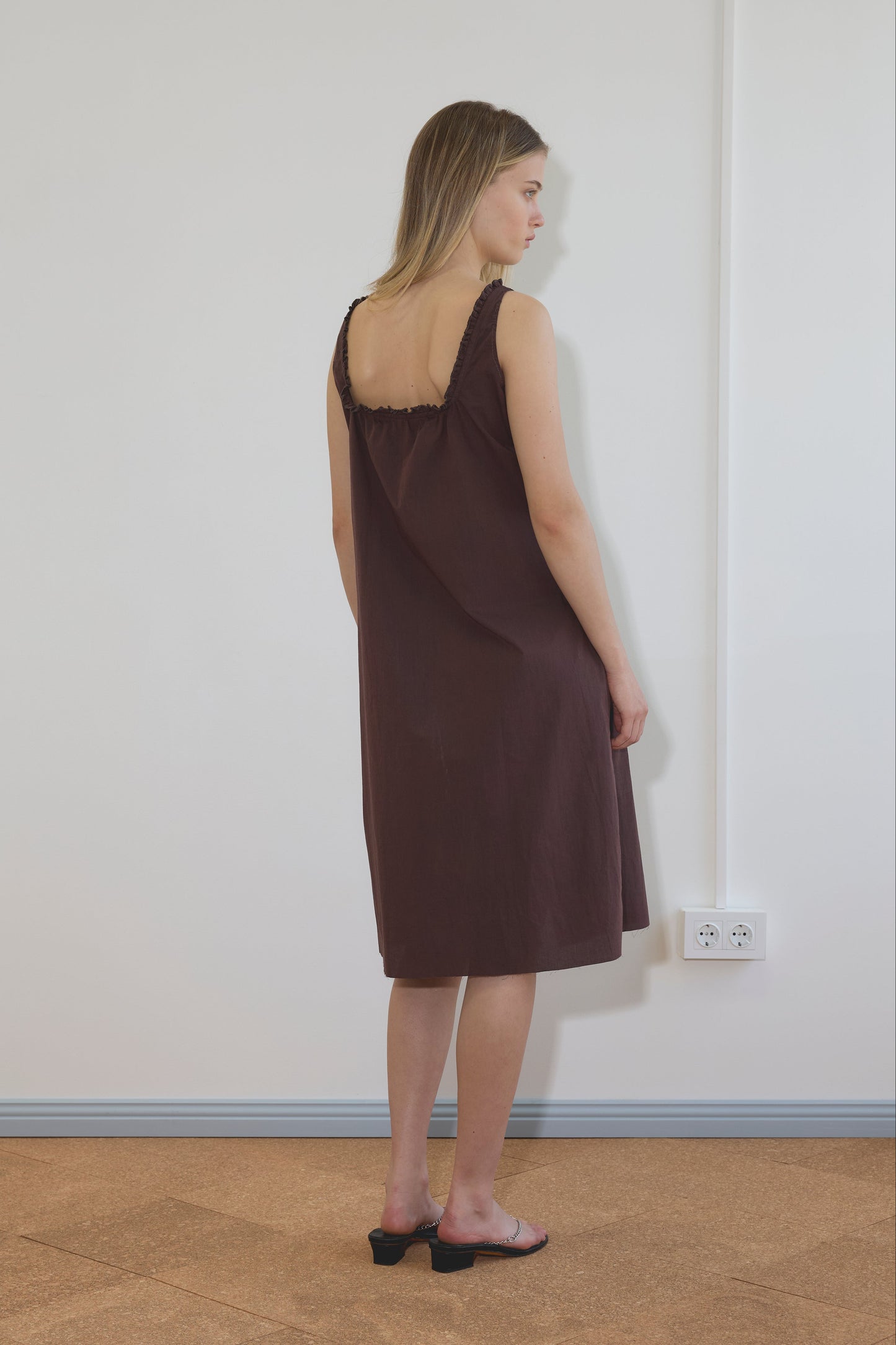 A woman stands indoors in the Button Sweep Dress - Umber, a loose, sleeveless dark brown dress with ruffled straps, paired with black heeled sandals. The setting features a plain white wall, blue baseboard, and electrical outlets.