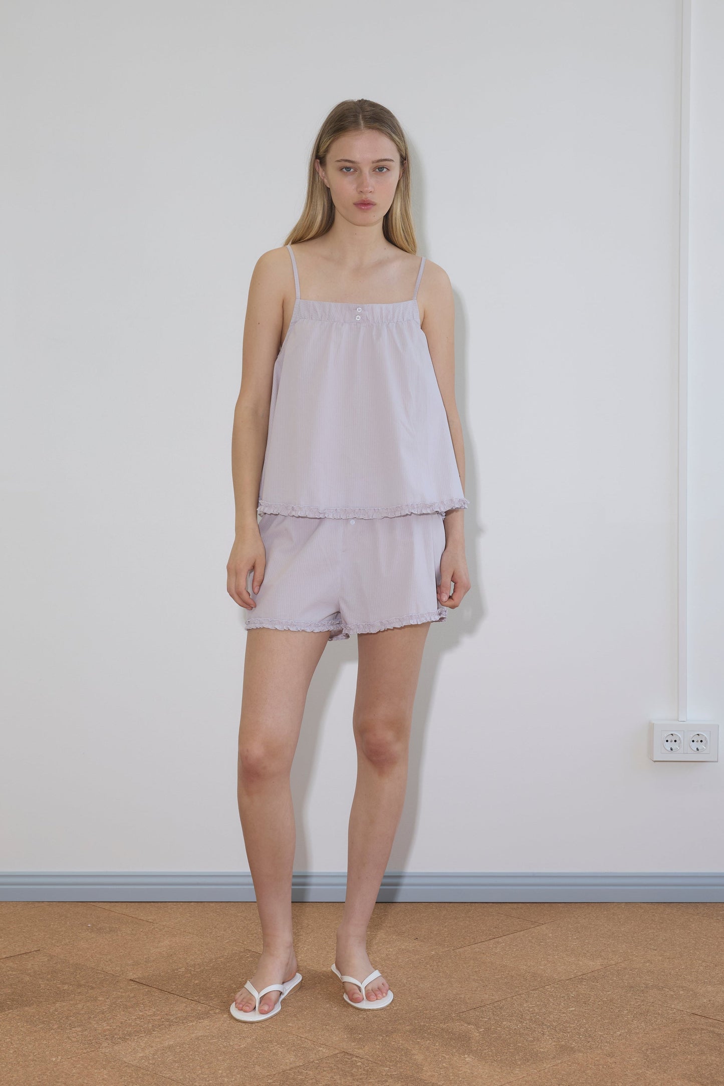 A woman stands indoors against a plain white wall, wearing the Yoke Set - Lilac Stripe with white sandals. She has long, straight hair and a neutral expression.