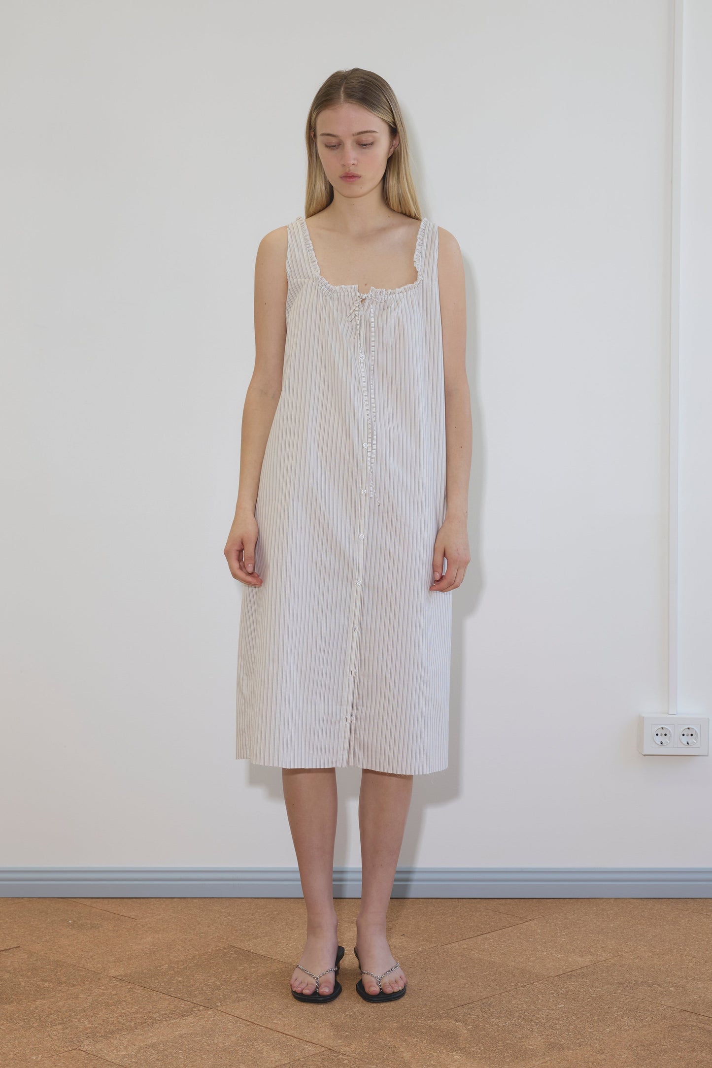 A woman with long, straight hair stands indoors against a white wall, wearing the knee-length Button Sweep Dress - Gull Stripe. The dress features thin straps, light stripes, buttons down the front, and is paired with sandals.