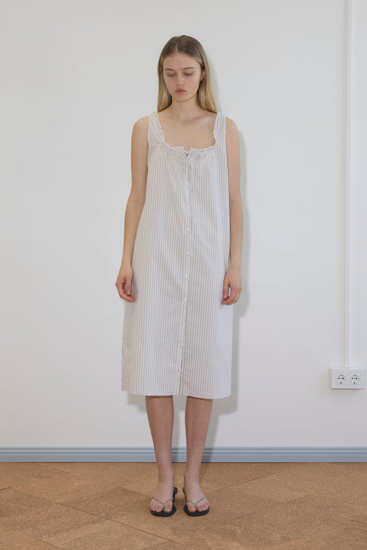A woman with long, straight hair stands indoors against a white wall, wearing the knee-length Button Sweep Dress - Gull Stripe. The dress features thin straps, light stripes, buttons down the front, and is paired with sandals.