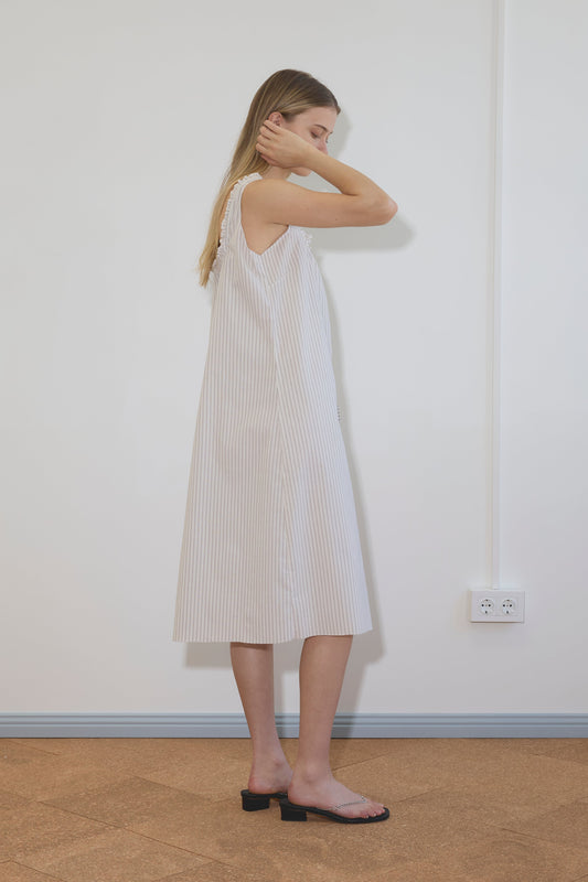 A woman stands sideways by a white wall, wearing the Button Sweep Dress - Gull Stripe, a loose, sleeveless organic cotton dress with light stripes, paired with black heeled sandals. She has long hair and touches her head. An electrical outlet appears on the wall.