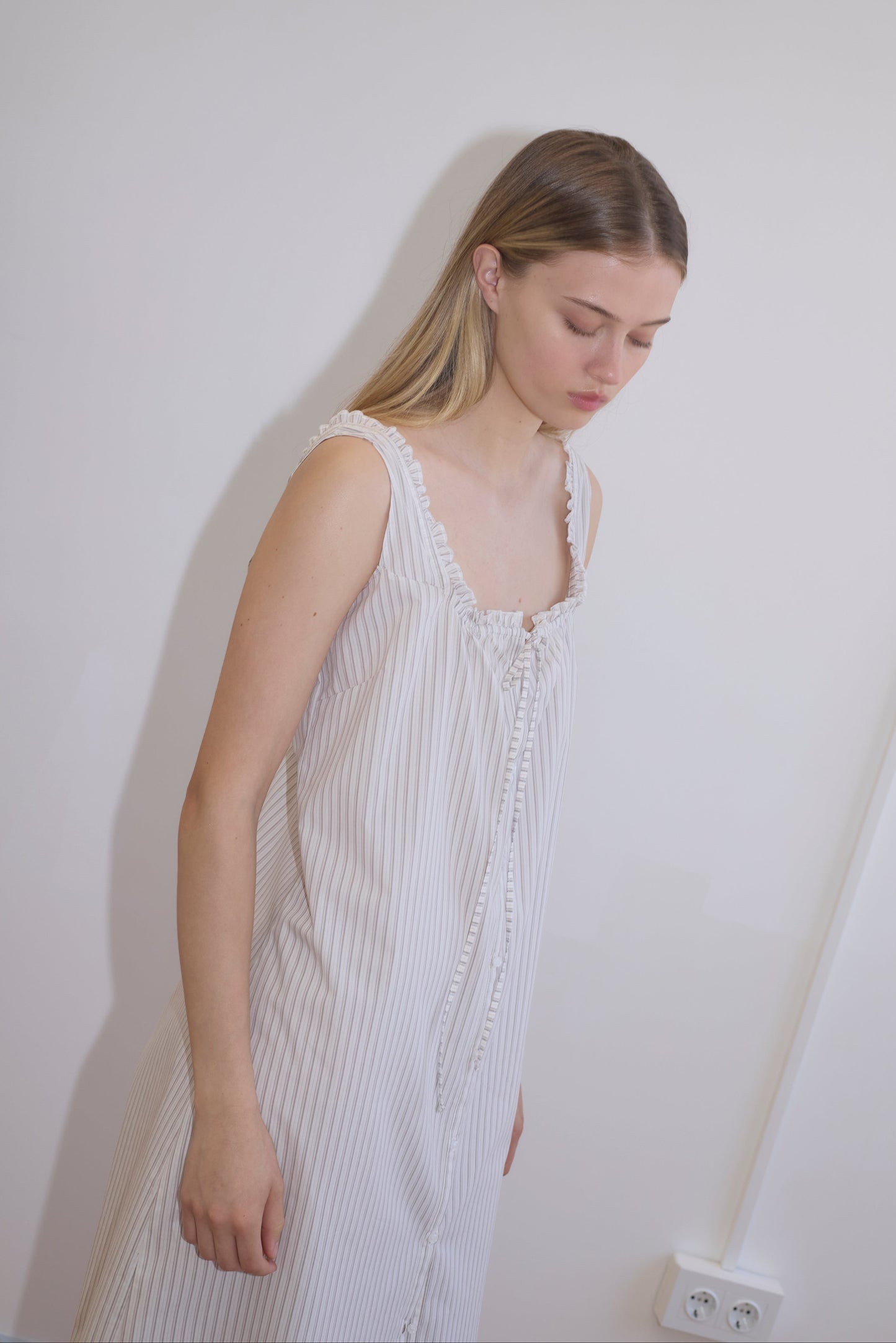 A young woman with long, straight blonde hair wears the Button Sweep Dress - Gull Stripe, a sleeveless white pinstriped organic cotton dress, standing against a plain white wall and looking downwards calmly.