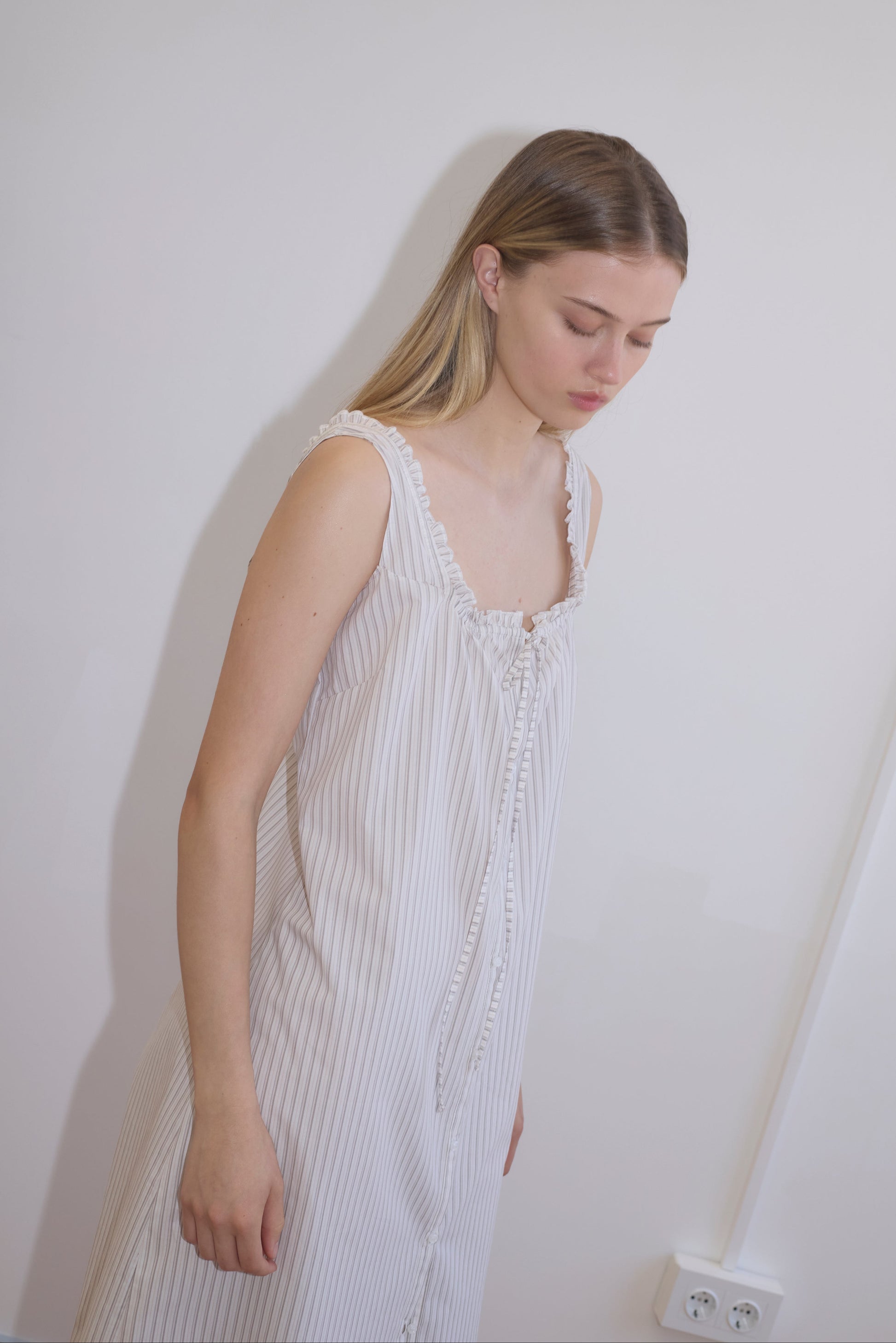 A young woman with long, straight blonde hair wears the Button Sweep Dress - Gull Stripe, a sleeveless white pinstriped organic cotton dress, standing against a plain white wall and looking downwards calmly.