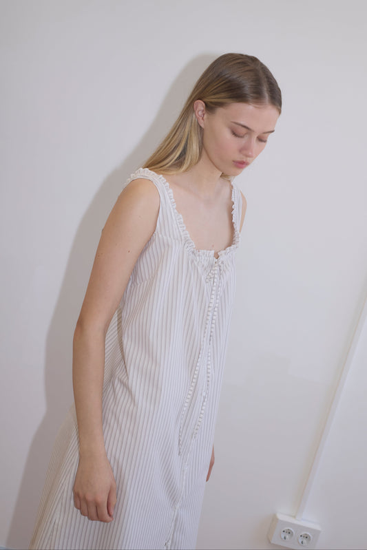 A young woman with long, straight blonde hair wears the Button Sweep Dress - Gull Stripe, a sleeveless white pinstriped organic cotton dress, standing against a plain white wall and looking downwards calmly.