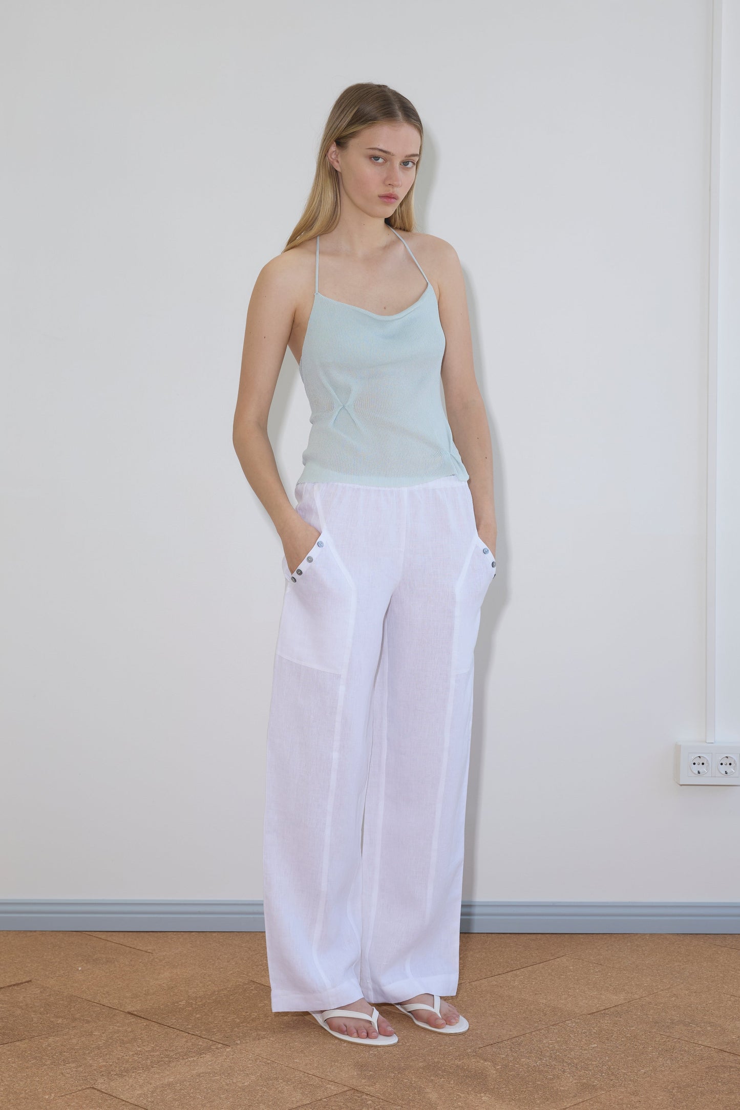 A woman stands indoors with hands in pockets, wearing a light blue spaghetti strap top, Deiji Studios Angle Pocket Pant in white, and white sandals. She has long blonde hair and a neutral expression against a plain light wall.