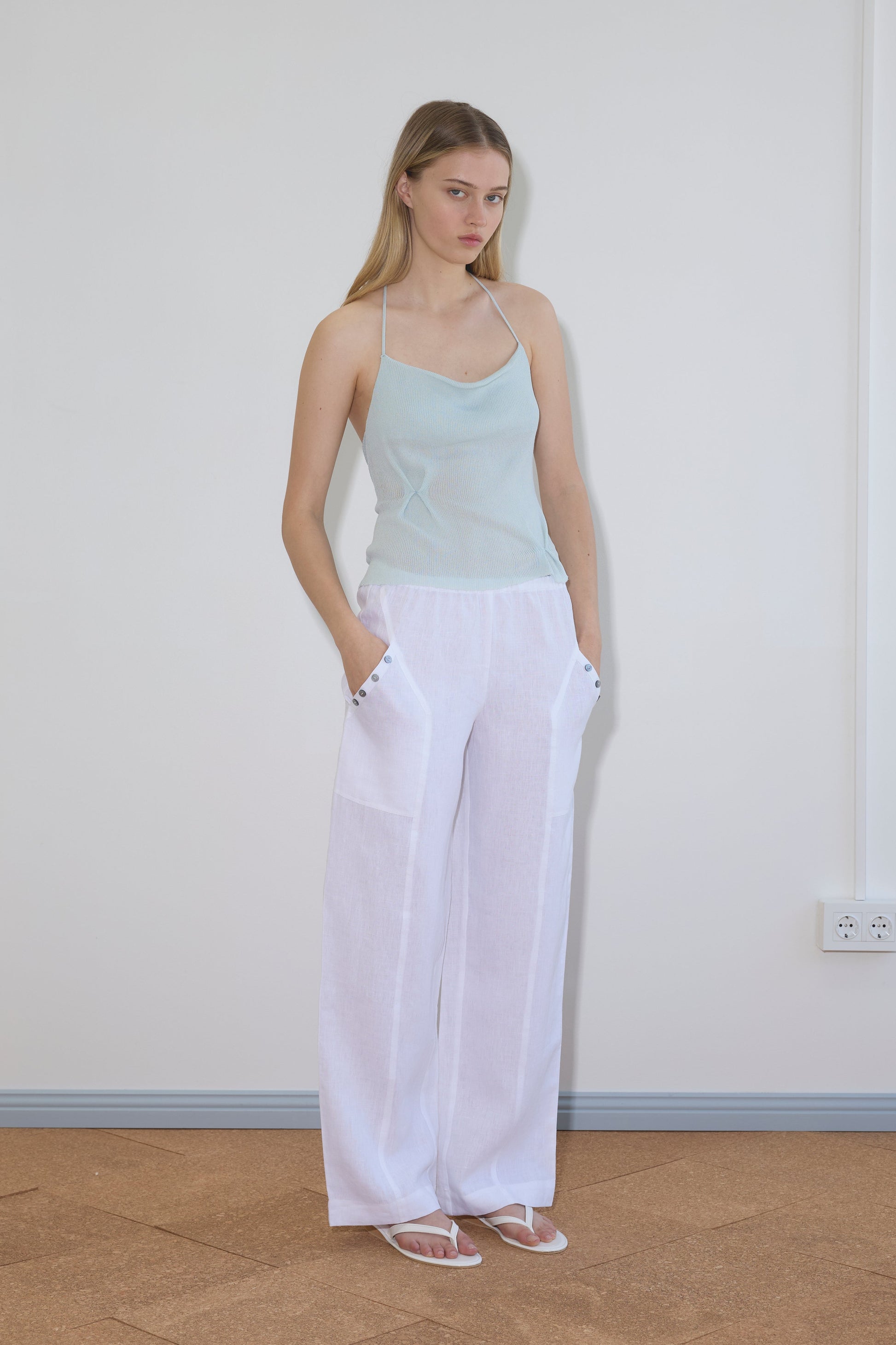 A woman stands indoors with hands in pockets, wearing a light blue spaghetti strap top, Deiji Studios Angle Pocket Pant in white, and white sandals. She has long blonde hair and a neutral expression against a plain light wall.