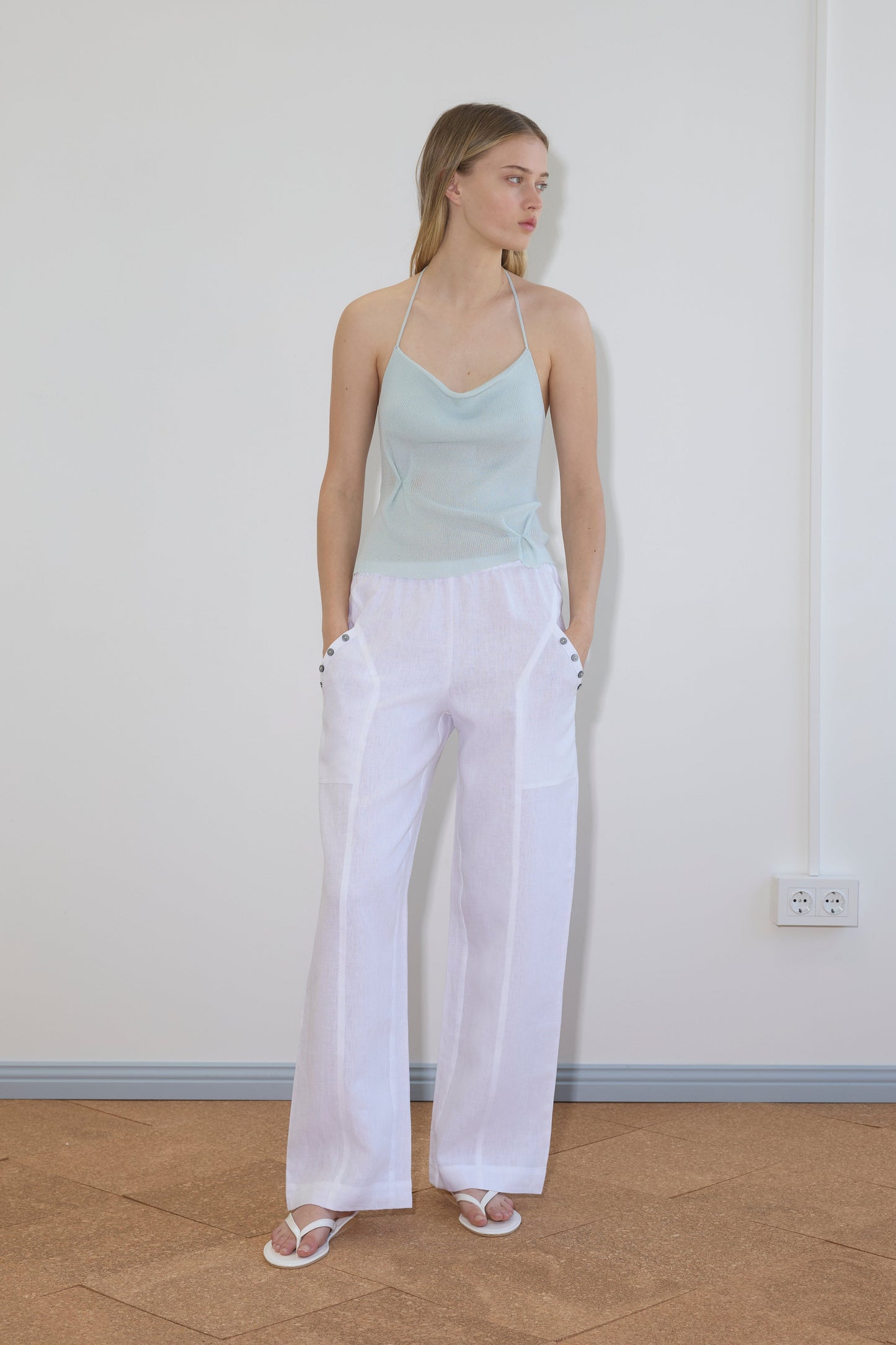 A woman with light brown hair stands indoors against a white wall, wearing the Angle Pocket Pant in white with pockets, a light blue halter top, and white sandals. Her hands rest in her pockets.