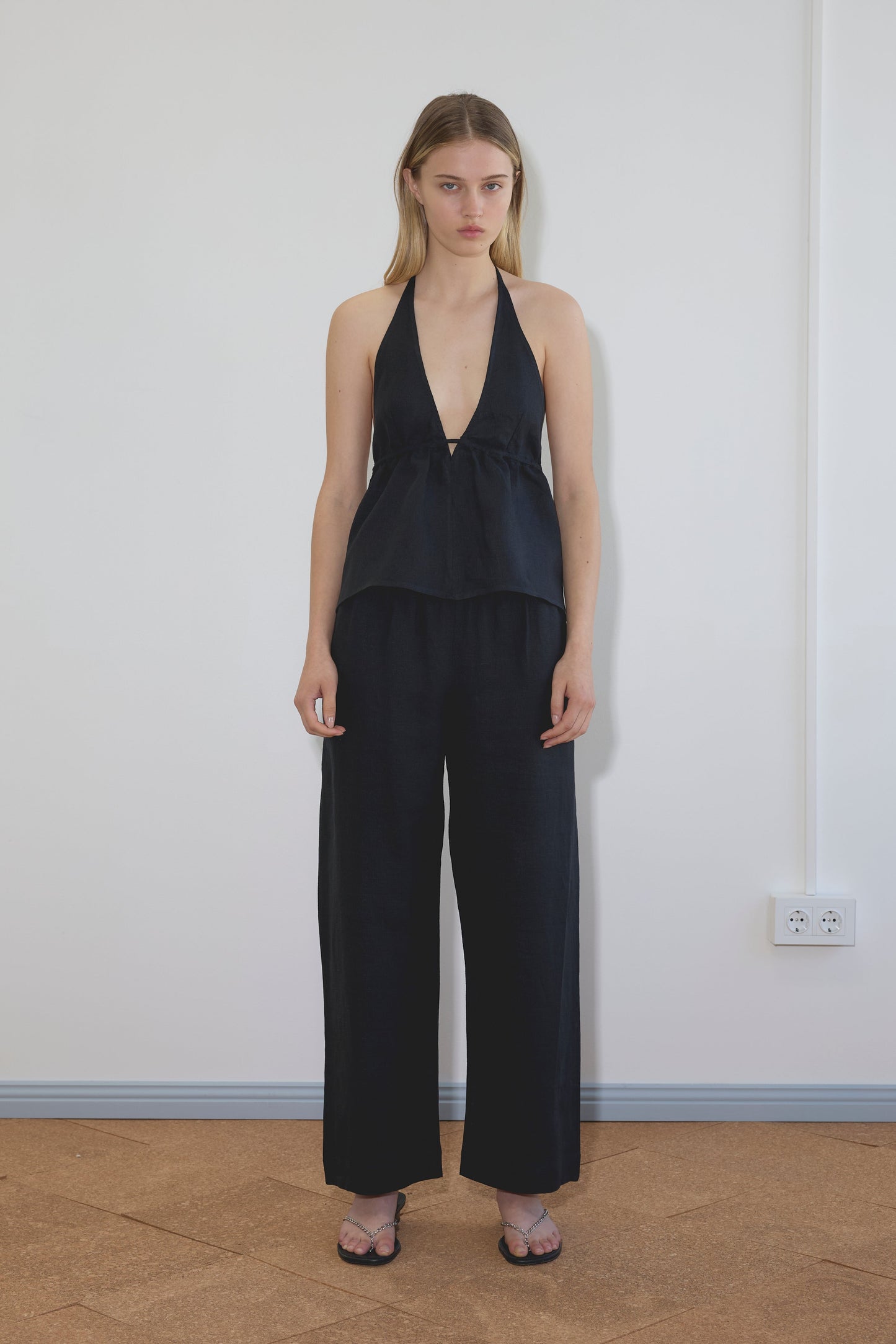 A woman stands indoors against a white wall, wearing Deiji Studios’ Linen Button Halter in black with loose black wide-leg pants. She has straight, mid-length hair and is wearing black sandals.