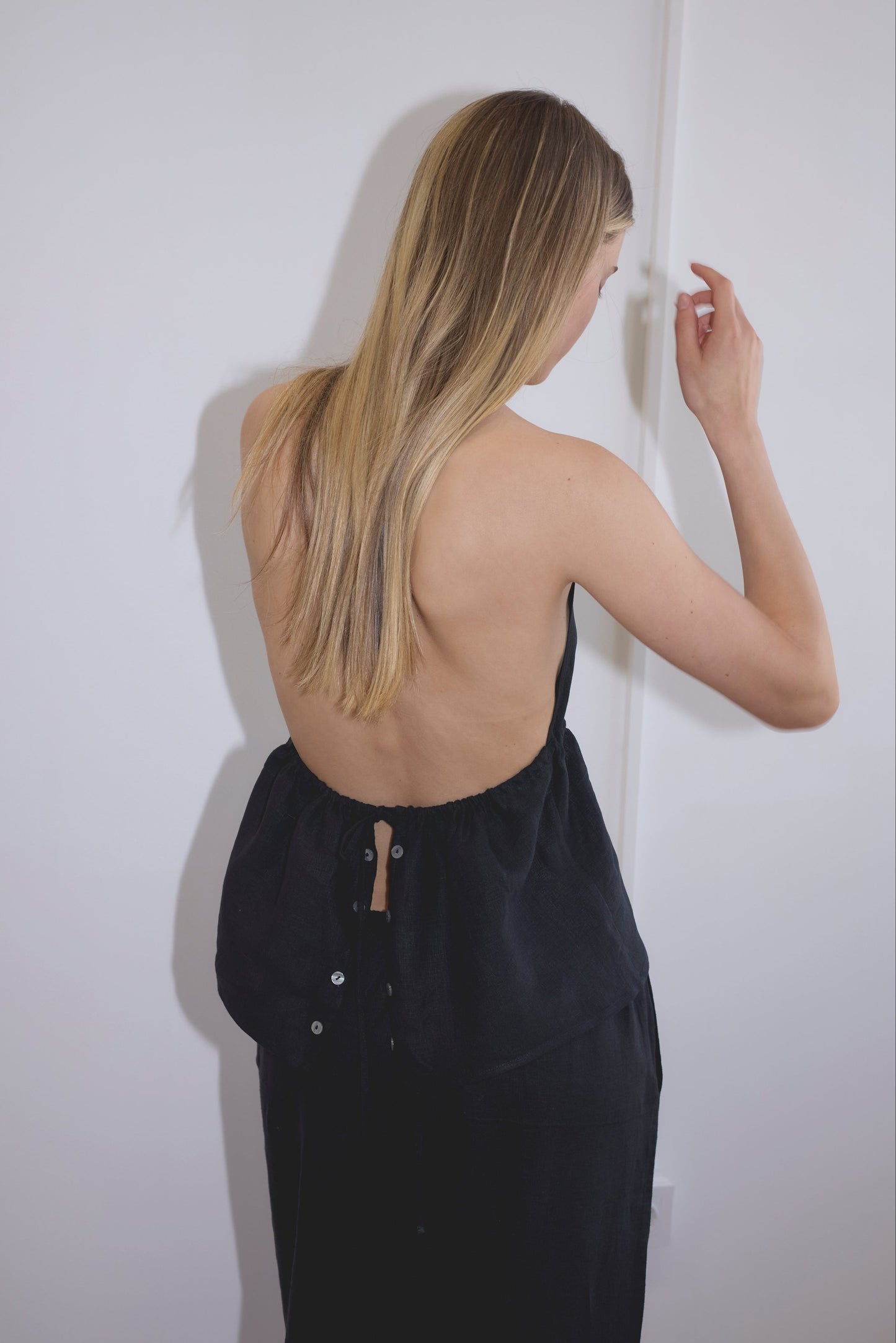 A woman with long blonde hair stands with her back to the camera, wearing a black Linen Button Halter by Deiji Studios and dark pants, in a simple white room.
