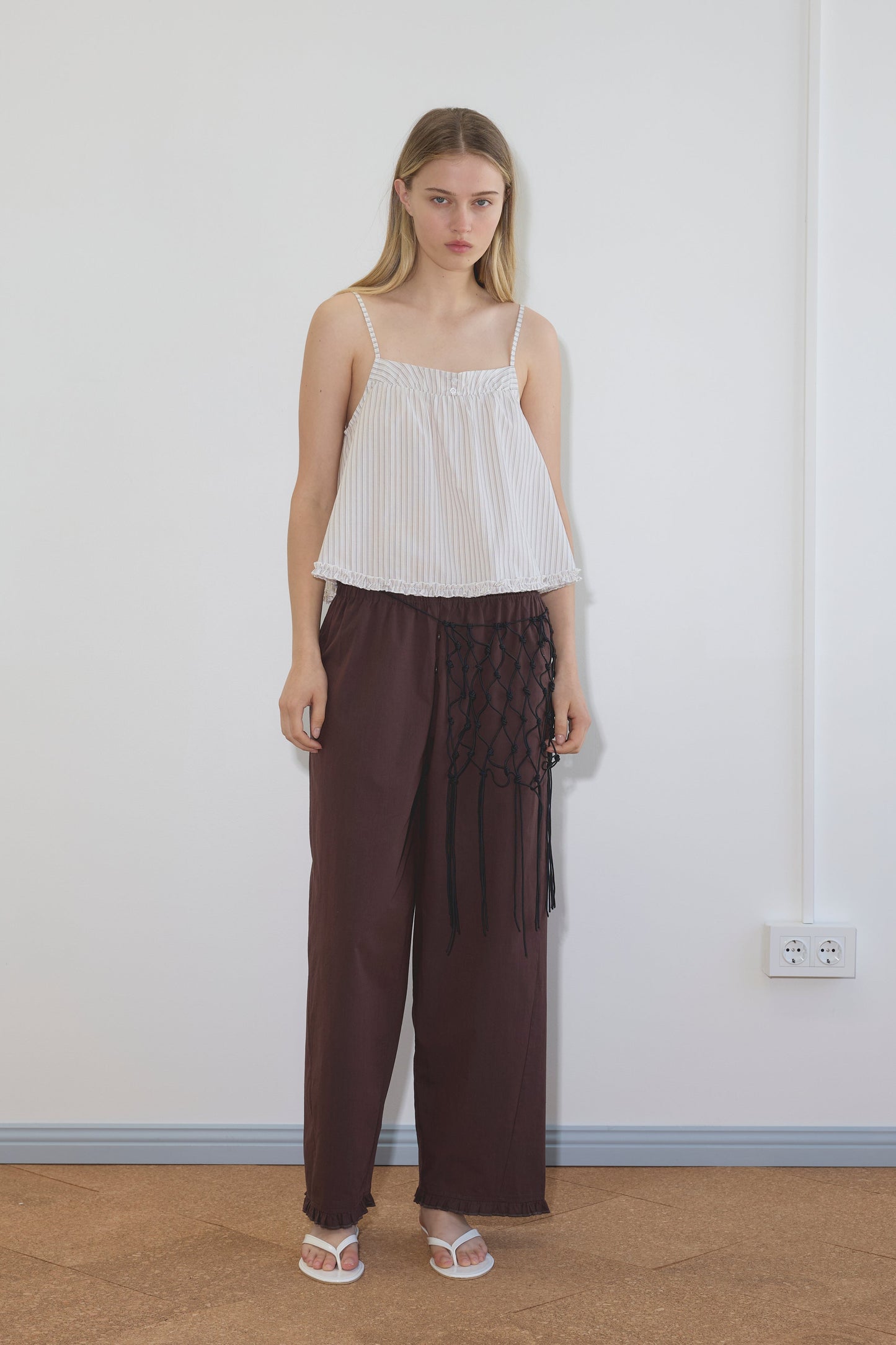 A woman with straight blonde hair stands indoors against a white wall, wearing a loose-fitting Deiji Studios Yoke Top in Gull Stripe, wide-legged maroon pants with black netted detail on one side, and white sandals.