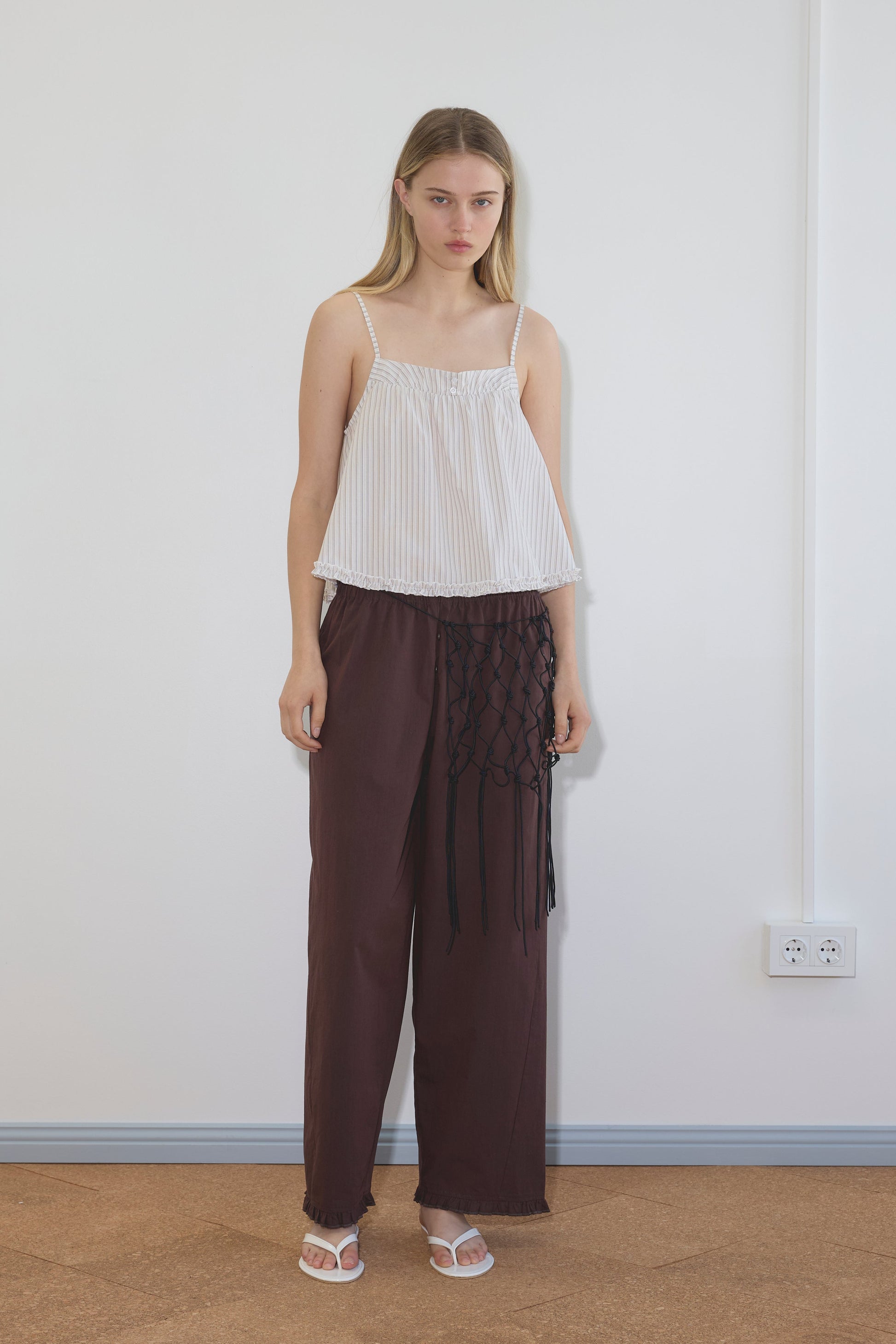 A woman with straight blonde hair stands indoors against a white wall, wearing a loose-fitting Deiji Studios Yoke Top in Gull Stripe, wide-legged maroon pants with black netted detail on one side, and white sandals.