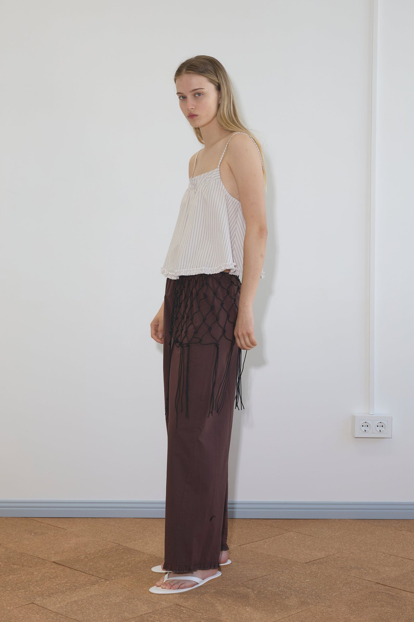 A woman stands indoors against a plain white wall, wearing the Yoke Top - Gull Stripe by Deiji Studios, dark brown pants with a black net overlay, and white sandals. She looks toward the camera with a neutral expression.