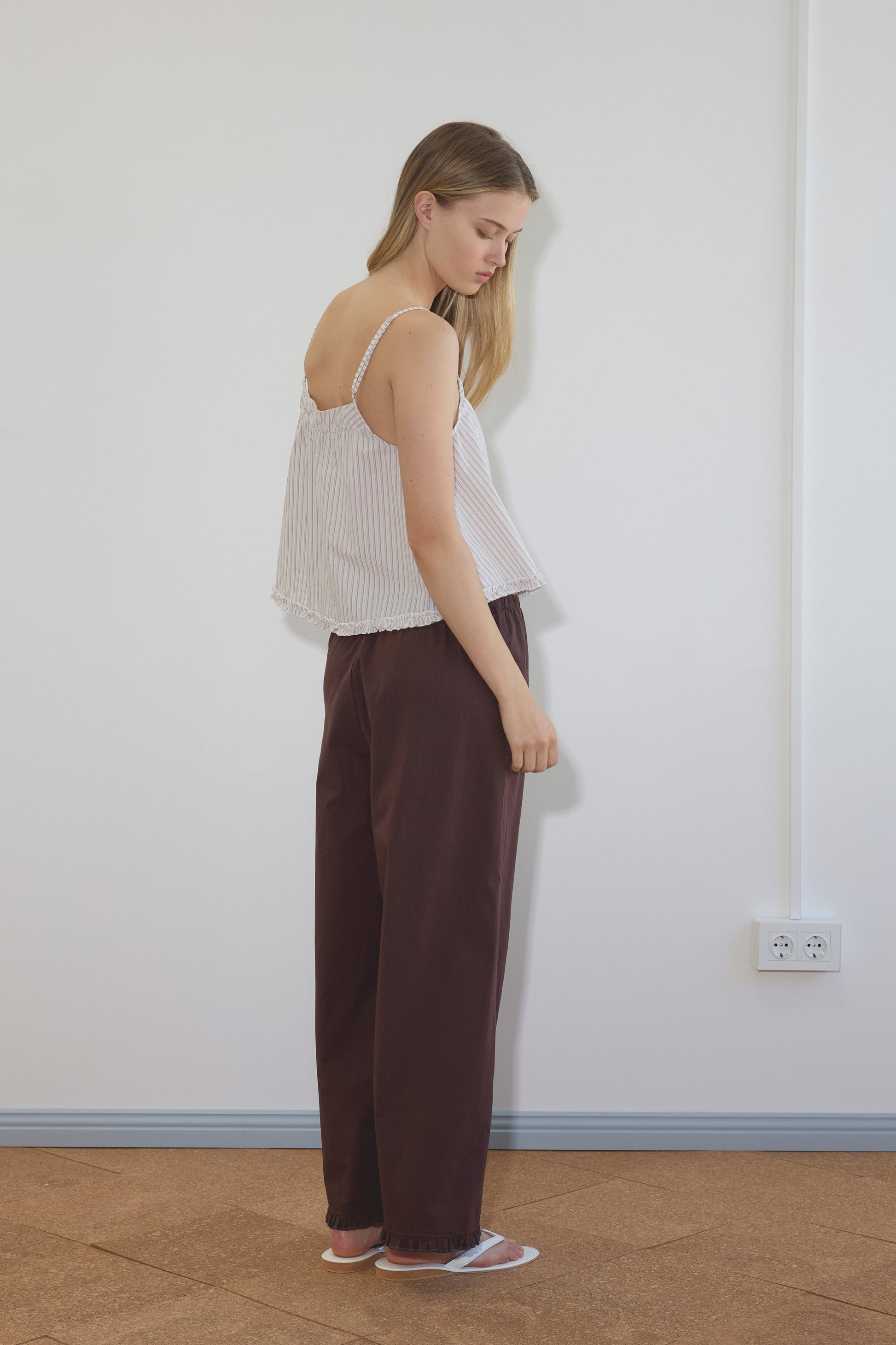 A woman with long blonde hair stands sideways in a minimal room, wearing Deiji Studios’ Yoke Top - Gull Stripe and wide-legged maroon pants, paired with white sandals.