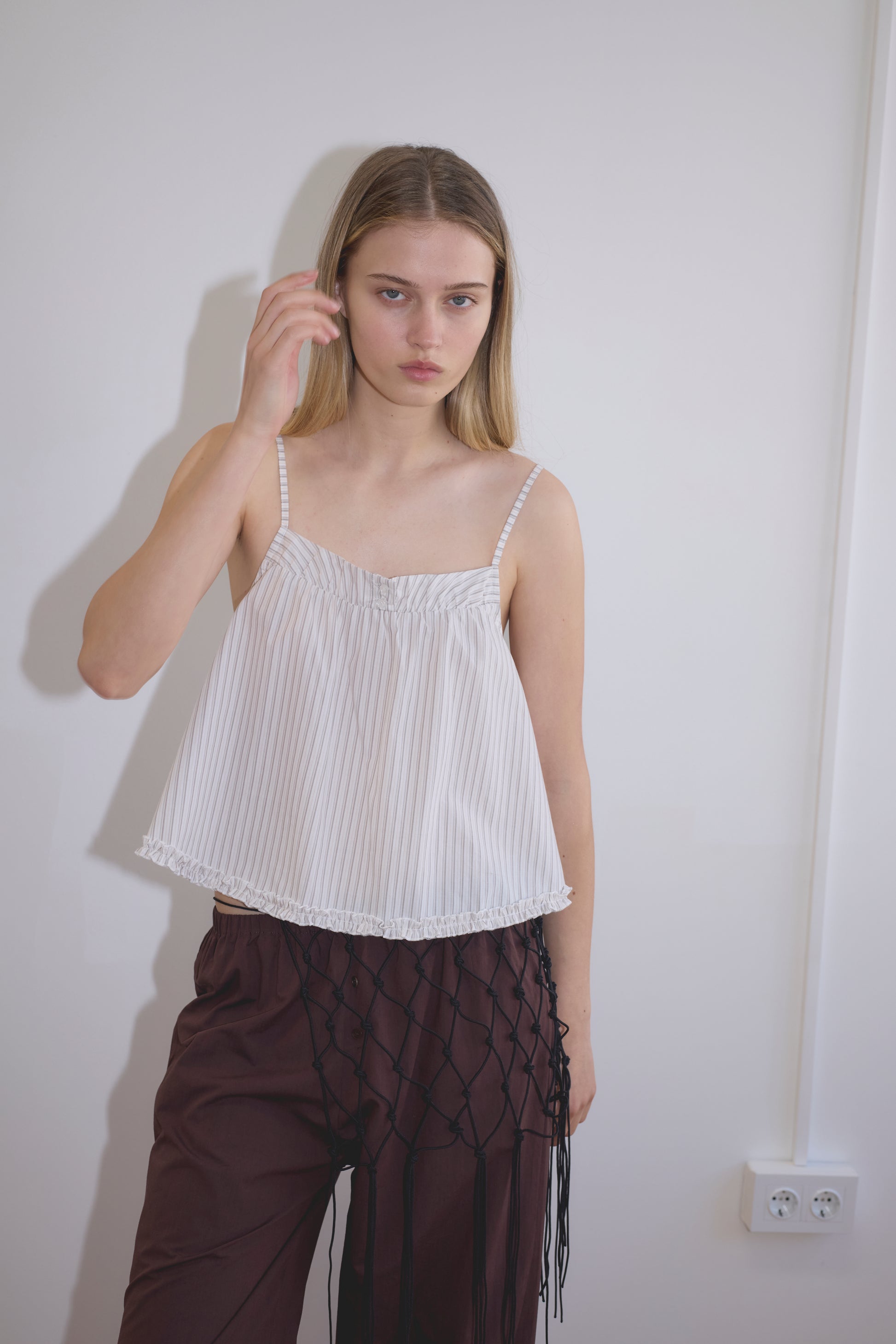 A young woman with straight blonde hair wears the Yoke Top - Gull Stripe from Deiji Studios and dark brown pants, standing against a plain white wall with a power outlet beside her.