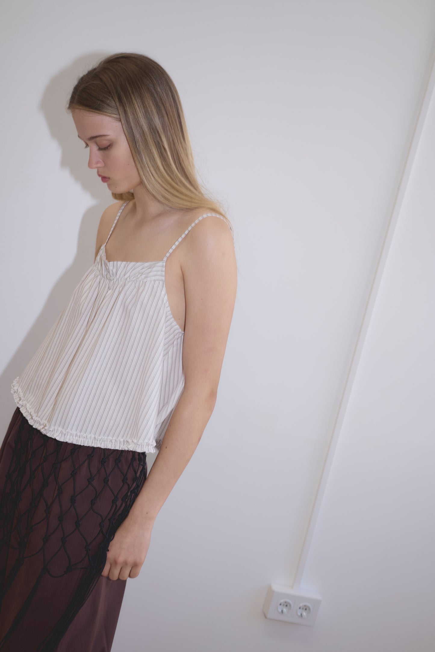 A woman with long, straight hair stands against a white wall, looking down. She wears the Yoke Top - Gull Stripe and a dark crisscross skirt. A power outlet is visible on the wall.