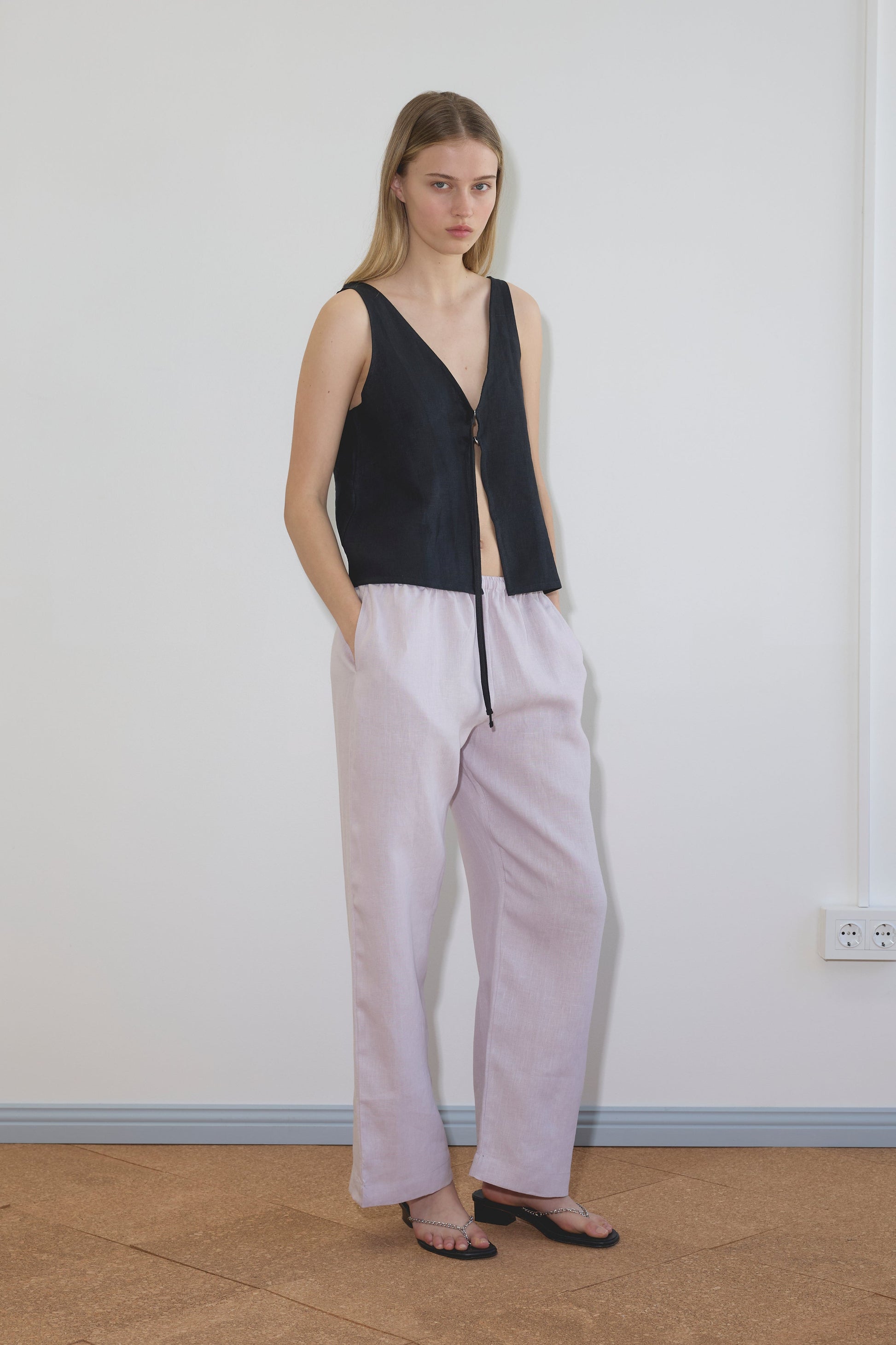 A woman stands indoors against a plain white wall, wearing the Two Part Top - Black by Deiji Studios with loose light pants and black sandals, her hands in her pockets.
