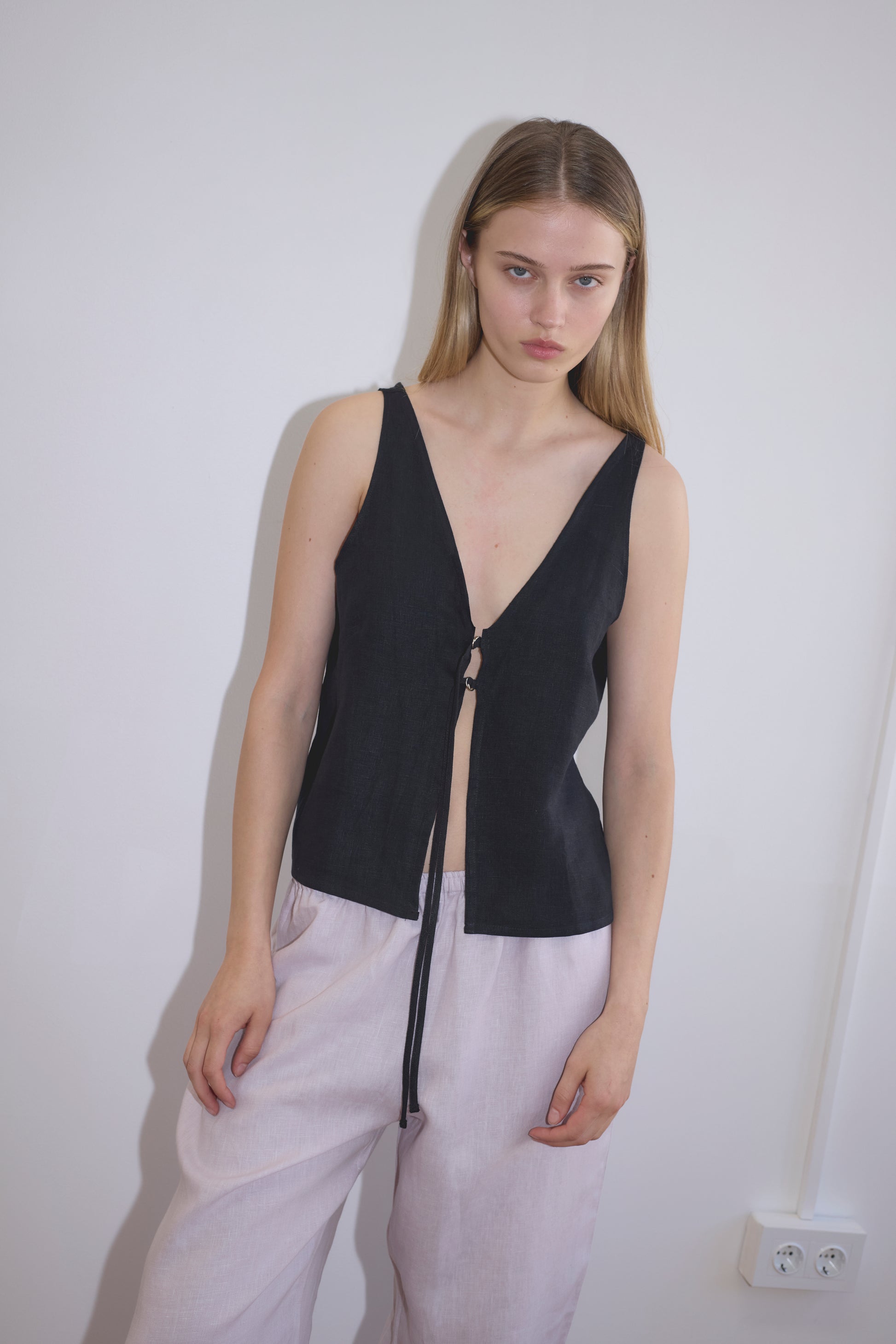 A young woman with long blonde hair stands against a white wall, wearing the Two Part Top - Black—a sleeveless, v-neck, tie-front style—paired with loose, light pants. She looks at the camera with a neutral expression, highlighting its versatile appeal.