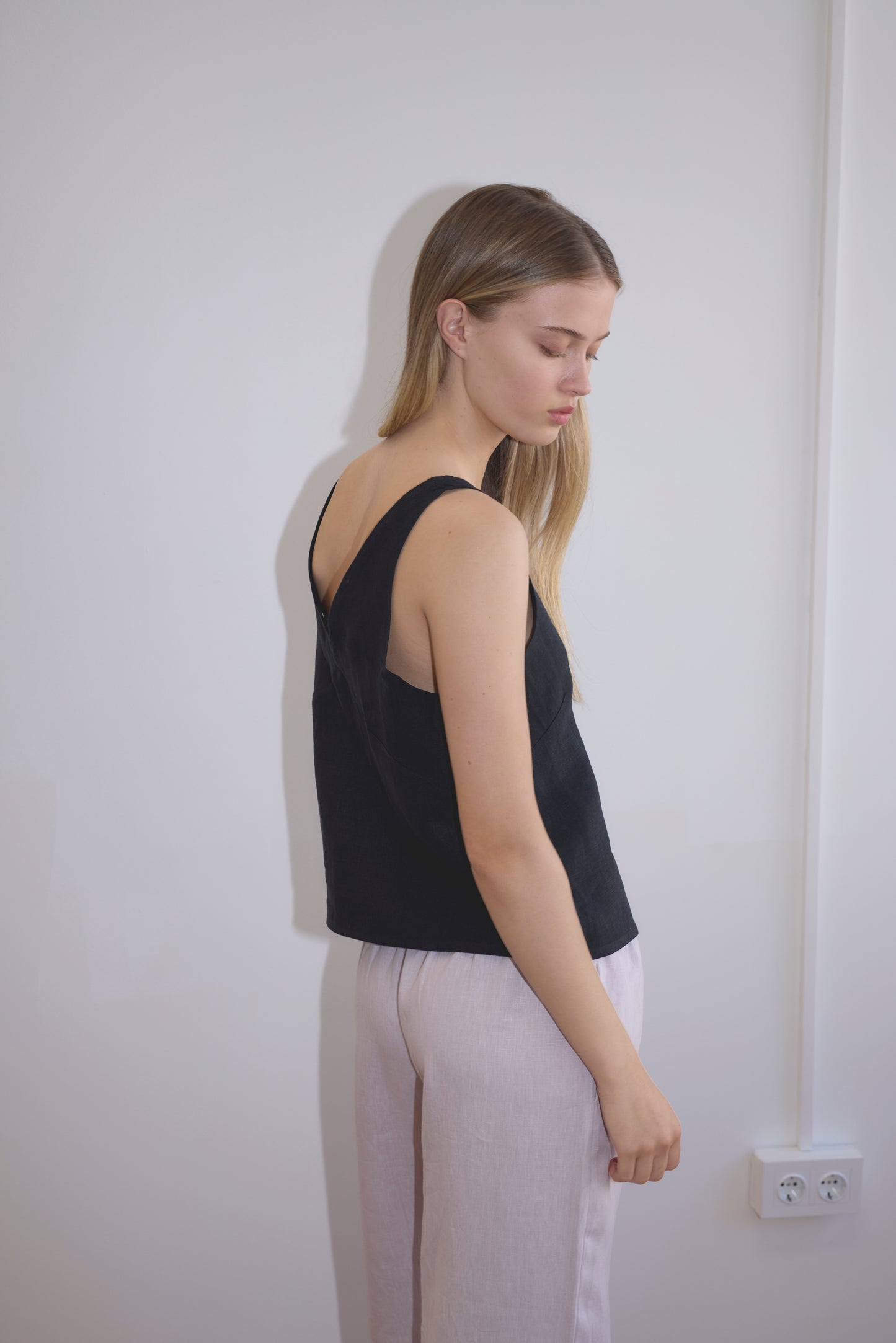 A woman with long blonde hair stands sideways by a plain white wall, wearing the Two Part Top - Black from Deiji Studios with light pants. She looks down with a neutral expression.