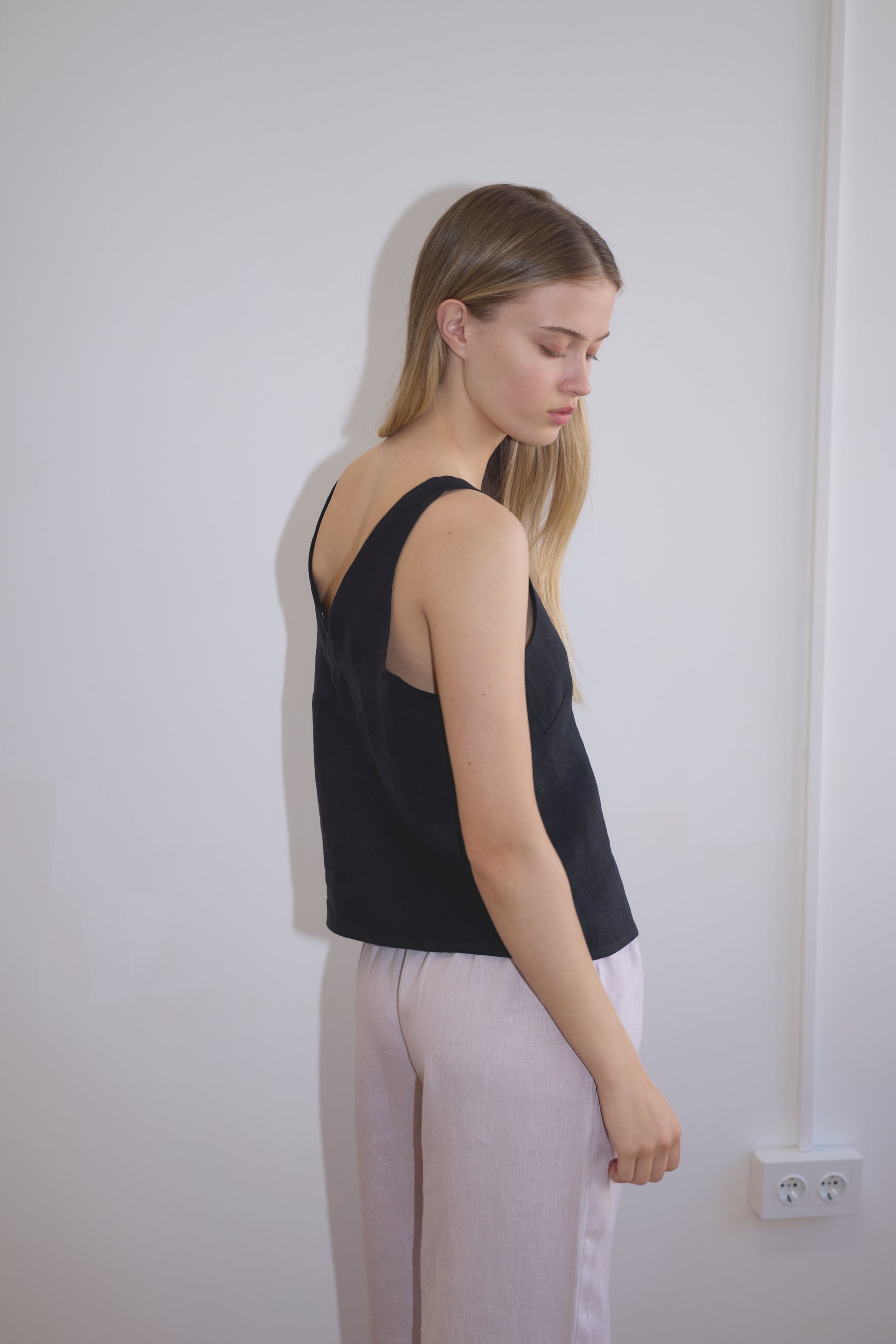 A woman with long blonde hair stands sideways by a plain white wall, wearing the Two Part Top - Black from Deiji Studios with light pants. She looks down with a neutral expression.