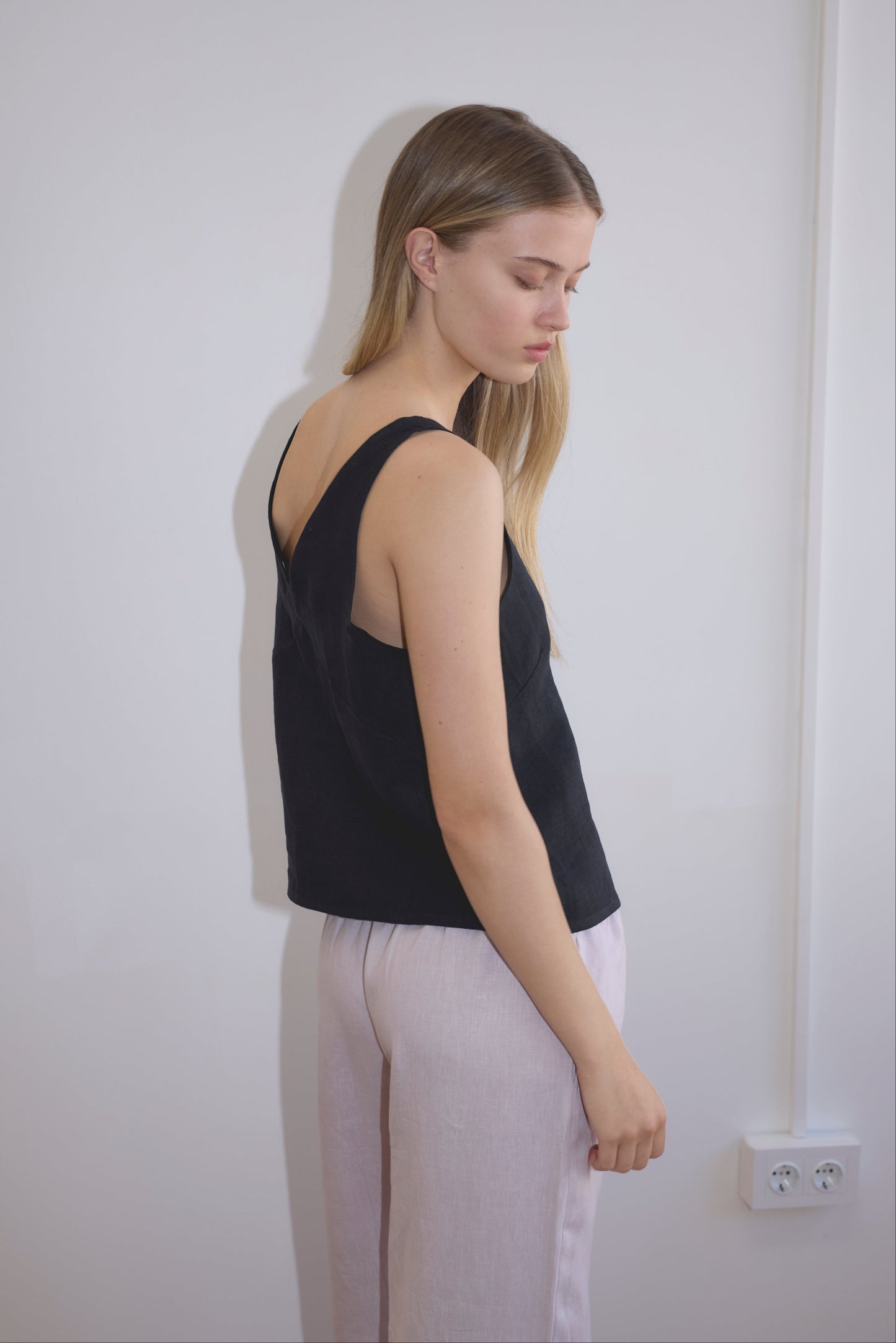 A woman with long, straight blonde hair stands sideways against a white wall, wearing a sleeveless black top and Linen Ease Trouser - Lilac. She looks downward with a calm expression. An electrical outlet is visible near the floor.