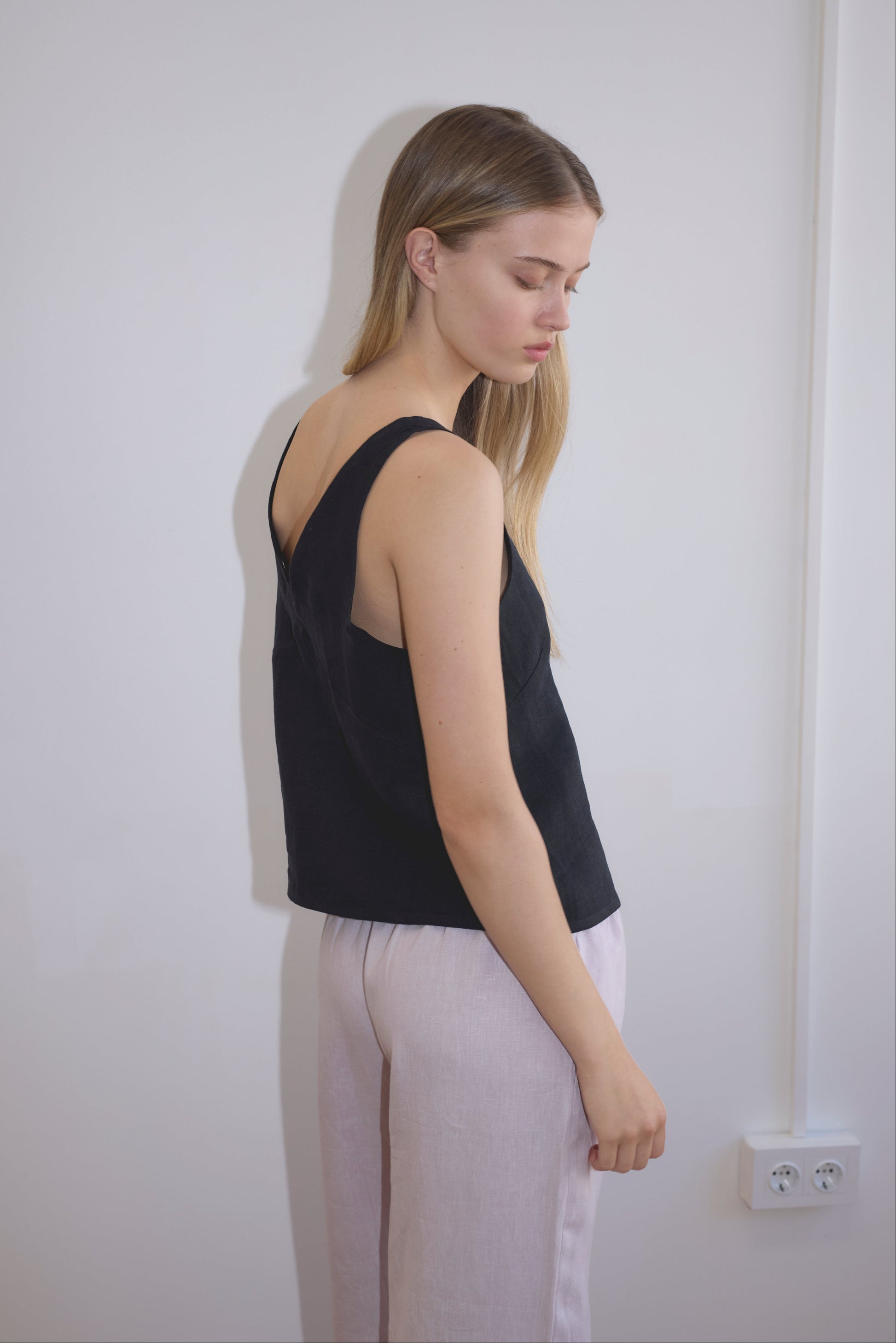 A woman with long, straight blonde hair stands sideways against a white wall, wearing a sleeveless black top and Linen Ease Trouser - Lilac. She looks downward with a calm expression. An electrical outlet is visible near the floor.