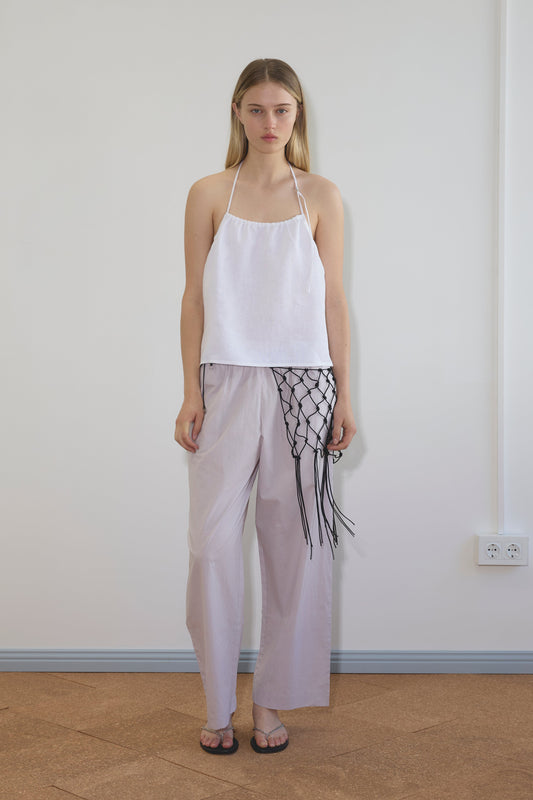 A woman stands indoors against a plain wall, wearing a white halter-top, Deiji Studios Ease Trouser in Lilac Stripe, black sandals, and a black netted accessory draped over her pants. A light brown floor and power outlet are visible.