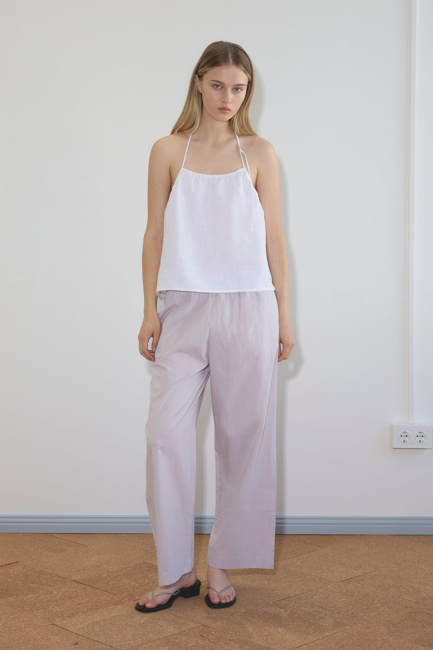 A woman stands indoors against a plain wall, wearing a white halter-style sleeveless top, Deiji Studios Ease Trouser in lilac stripe, and black flip-flops. She has straight blonde hair and a neutral expression.