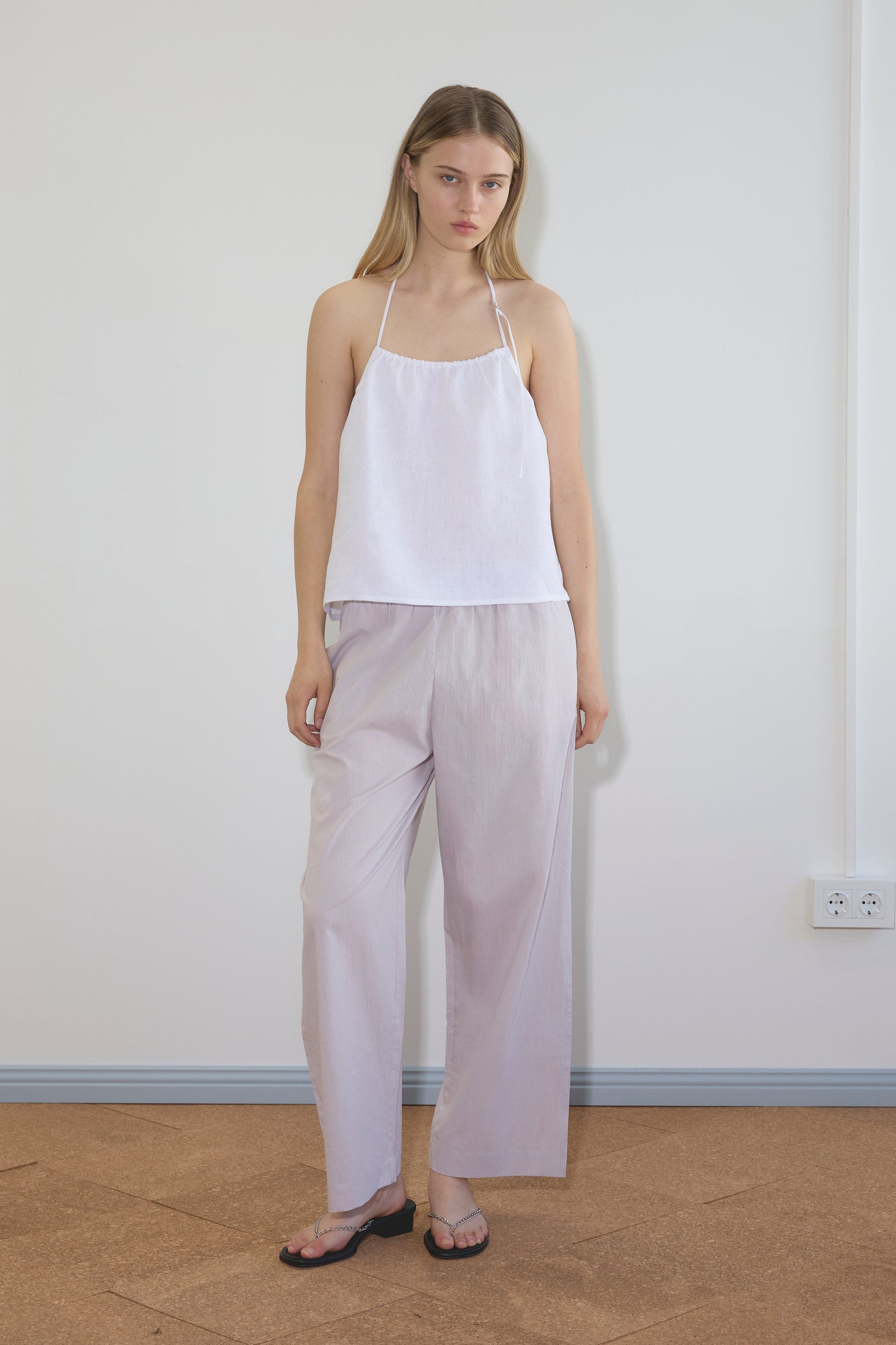 A woman stands indoors against a plain wall, wearing a white halter-style sleeveless top, Deiji Studios Ease Trouser in lilac stripe, and black flip-flops. She has straight blonde hair and a neutral expression.