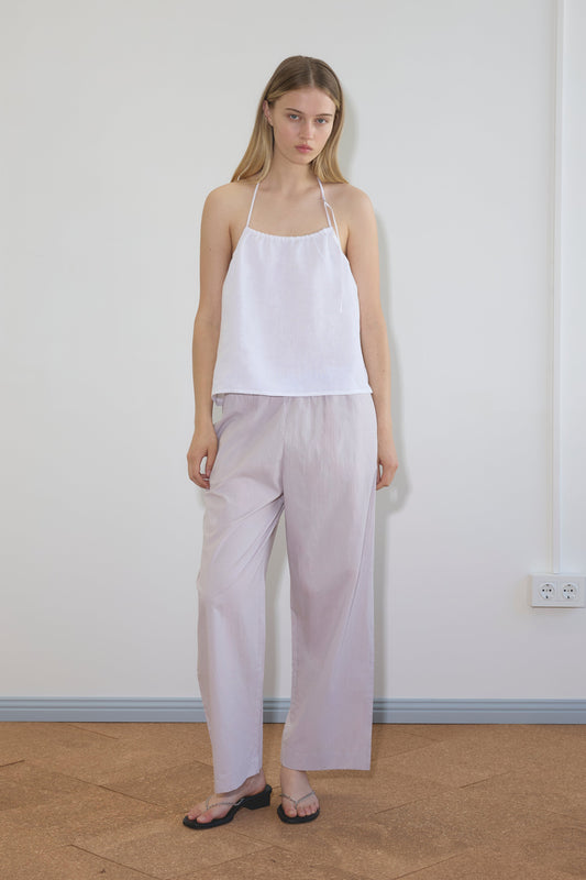 A woman stands indoors against a plain wall, wearing a white halter-style sleeveless top, Deiji Studios Ease Trouser in lilac stripe, and black flip-flops. She has straight blonde hair and a neutral expression.
