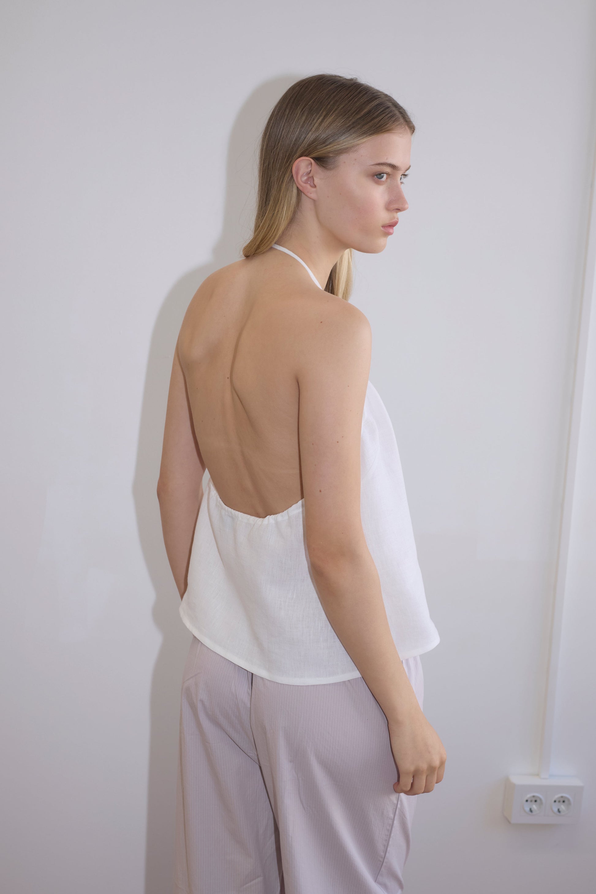 A young woman with long blonde hair stands sideways against a plain light wall, wearing the O Ring Halter - White and light-colored pants.