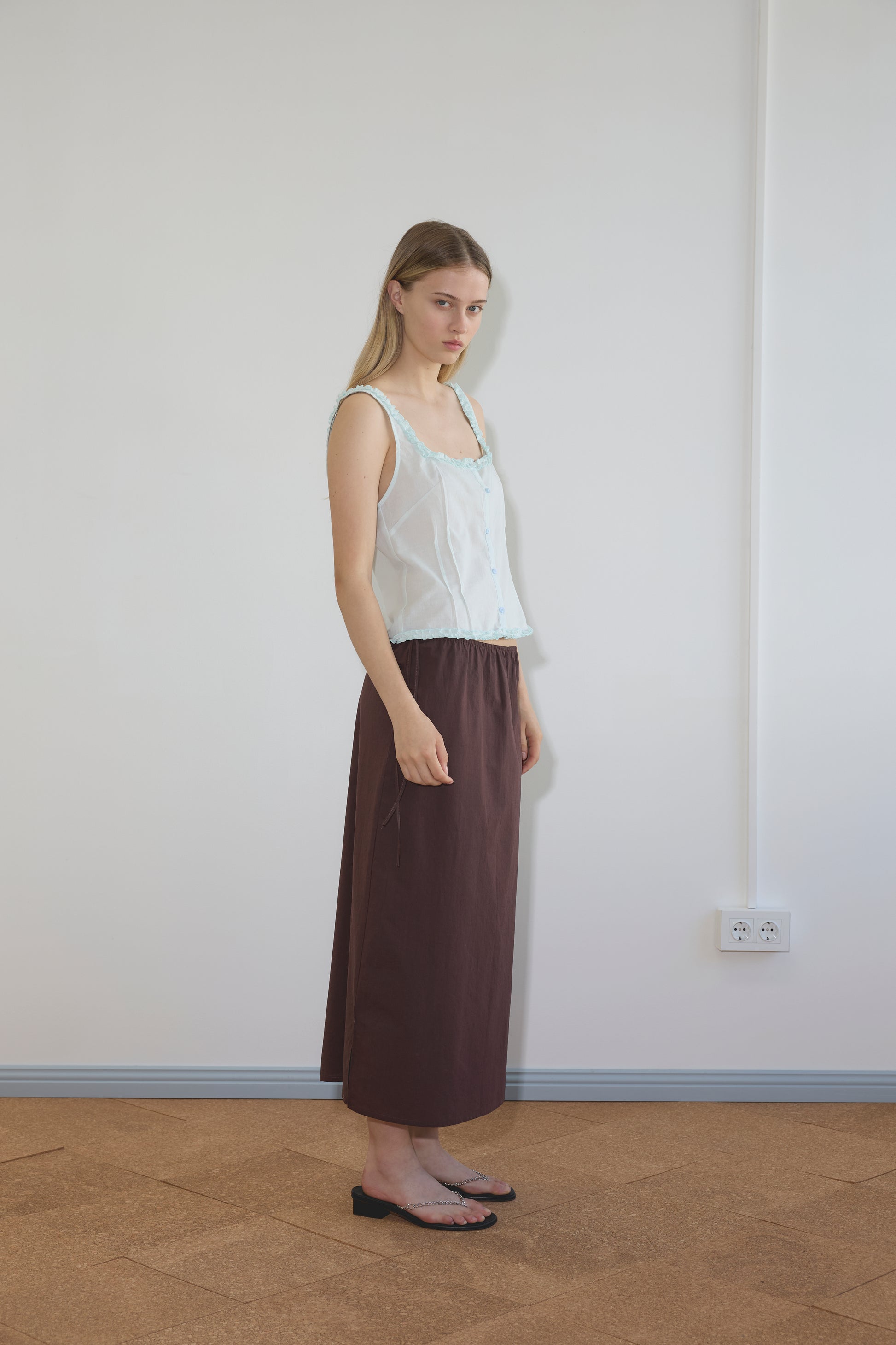 A young woman stands indoors against a white wall, wearing a sheer white sleeveless top, the Deiji Studios Mid Day Skirt - Umber, and black sandals. She faces slightly to the side with a neutral expression.