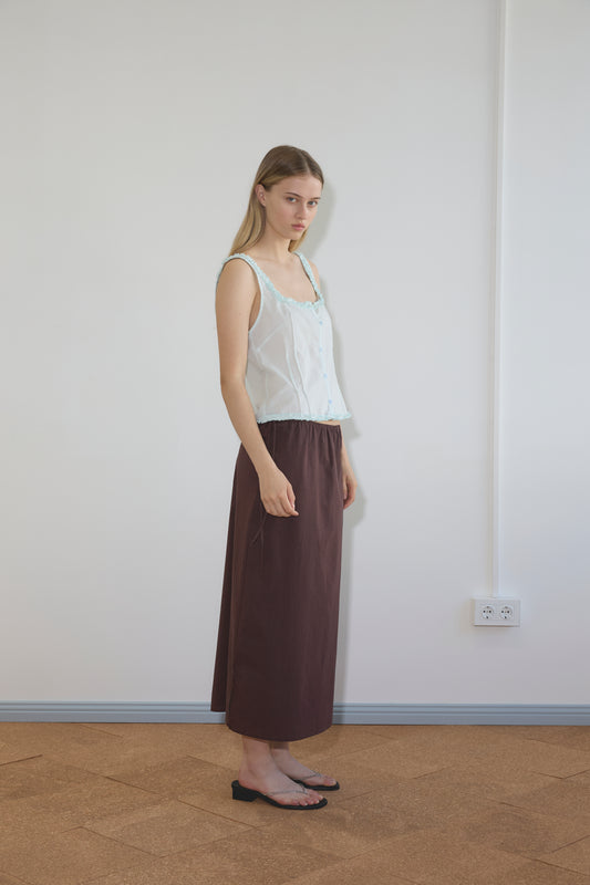 A young woman stands indoors against a white wall, wearing a sheer white sleeveless top, the Deiji Studios Mid Day Skirt - Umber, and black sandals. She faces slightly to the side with a neutral expression.