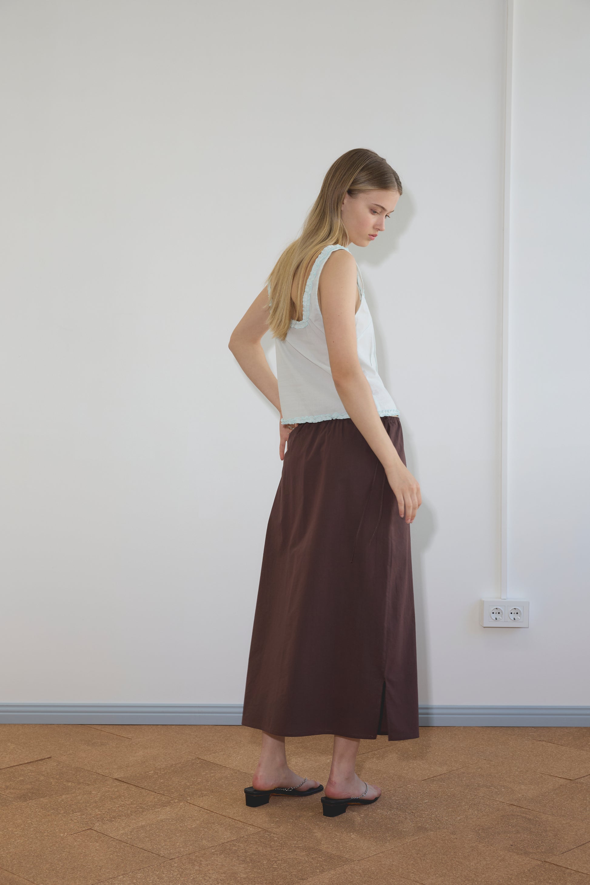 A woman with long blonde hair wearing a sleeveless light blue top and the Deiji Studios Mid Day Skirt – Umber stands sideways in a minimalist room with white walls and a cork floor.